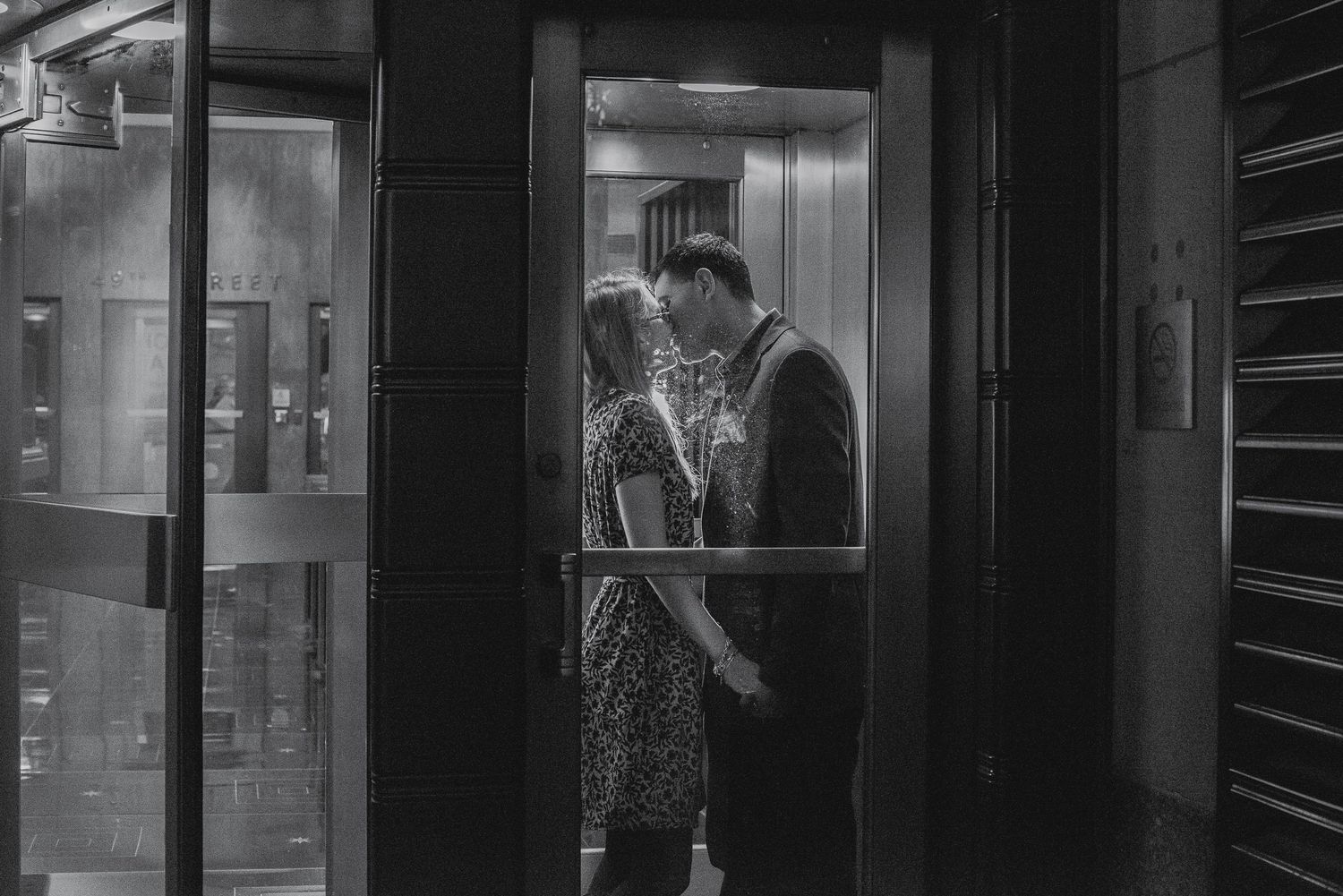 Black and white photo of a couple sharing an intimate moment inside a glass elevator at night.