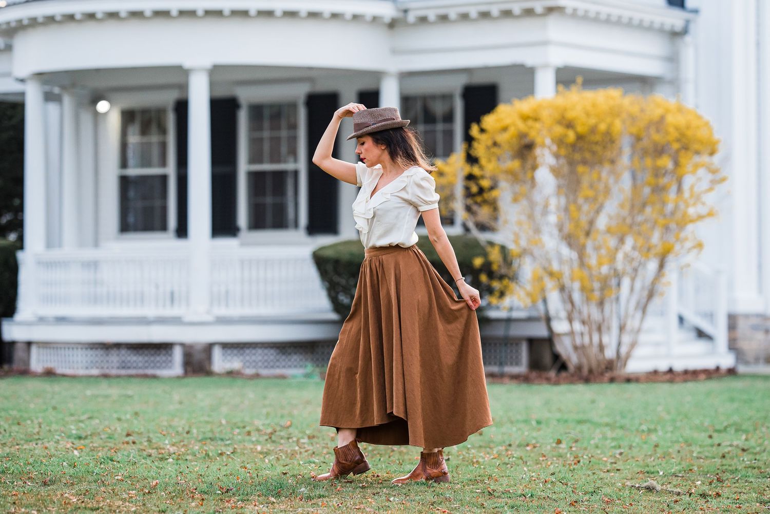 Stylish woman in brown midi skirt and hat posing on lawn in front of a white Victorian house.