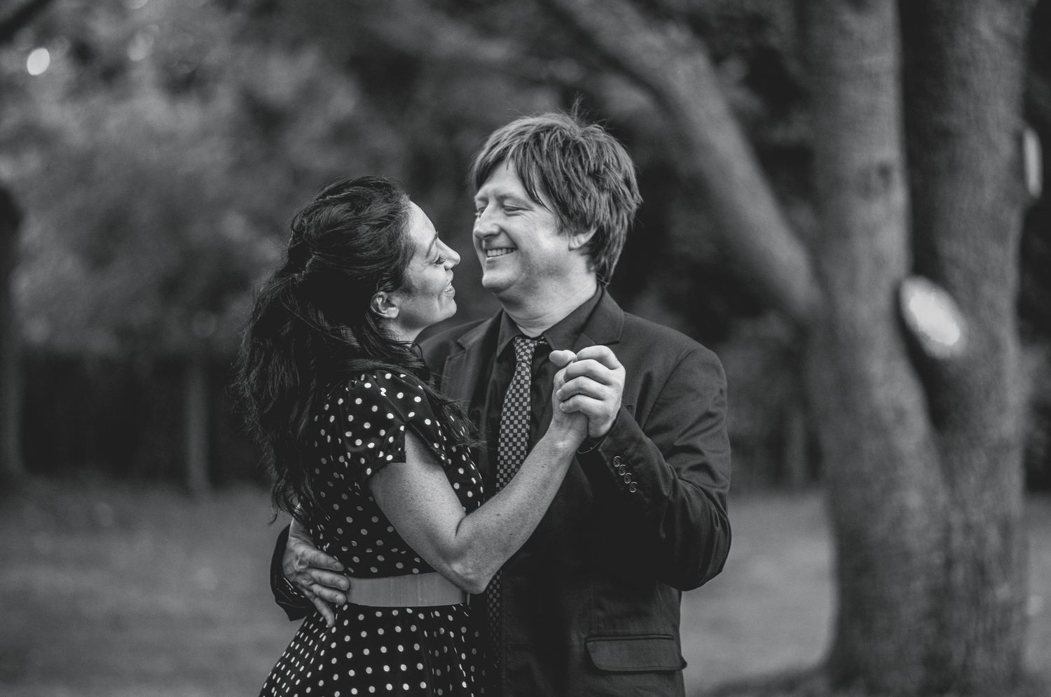 Black and white photo of a couple dancing together and smiling in a tree-lined outdoor setting.