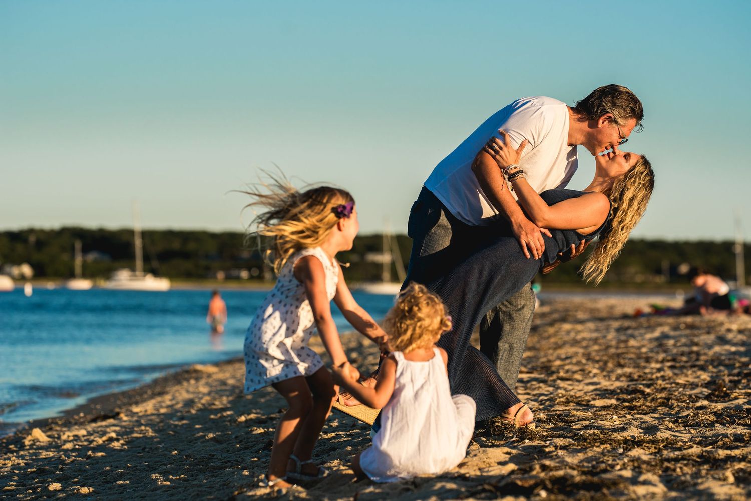 Family kissing on beach at sunset with two young girls in white dresses playing in the sand by the water.