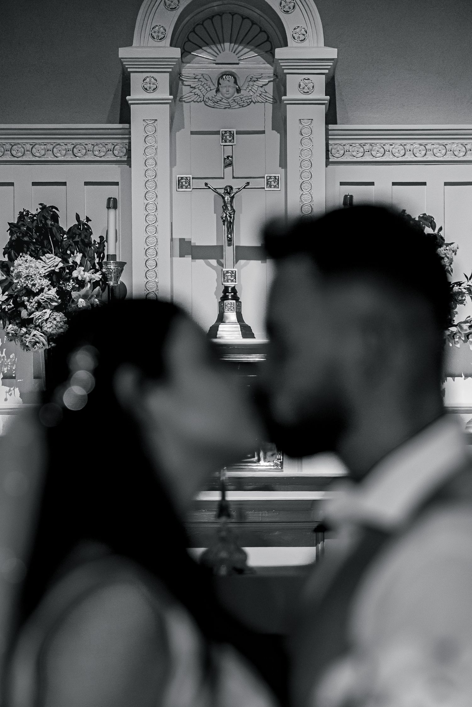 Black and white photo of a couple kissing at the altar of a church with a crucifix in the background.