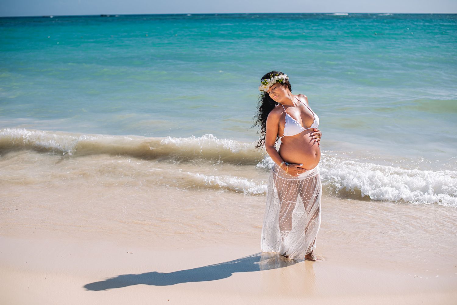 Pregnant woman in white sheer skirt and bikini top poses on tropical beach with turquoise ocean waves.