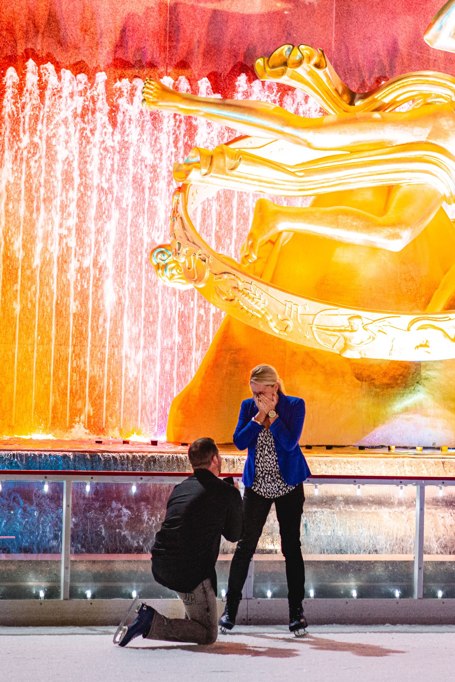 Man proposes on one knee at Rockefeller Center ice rink, woman reacts with surprise before golden Prometheus statue.