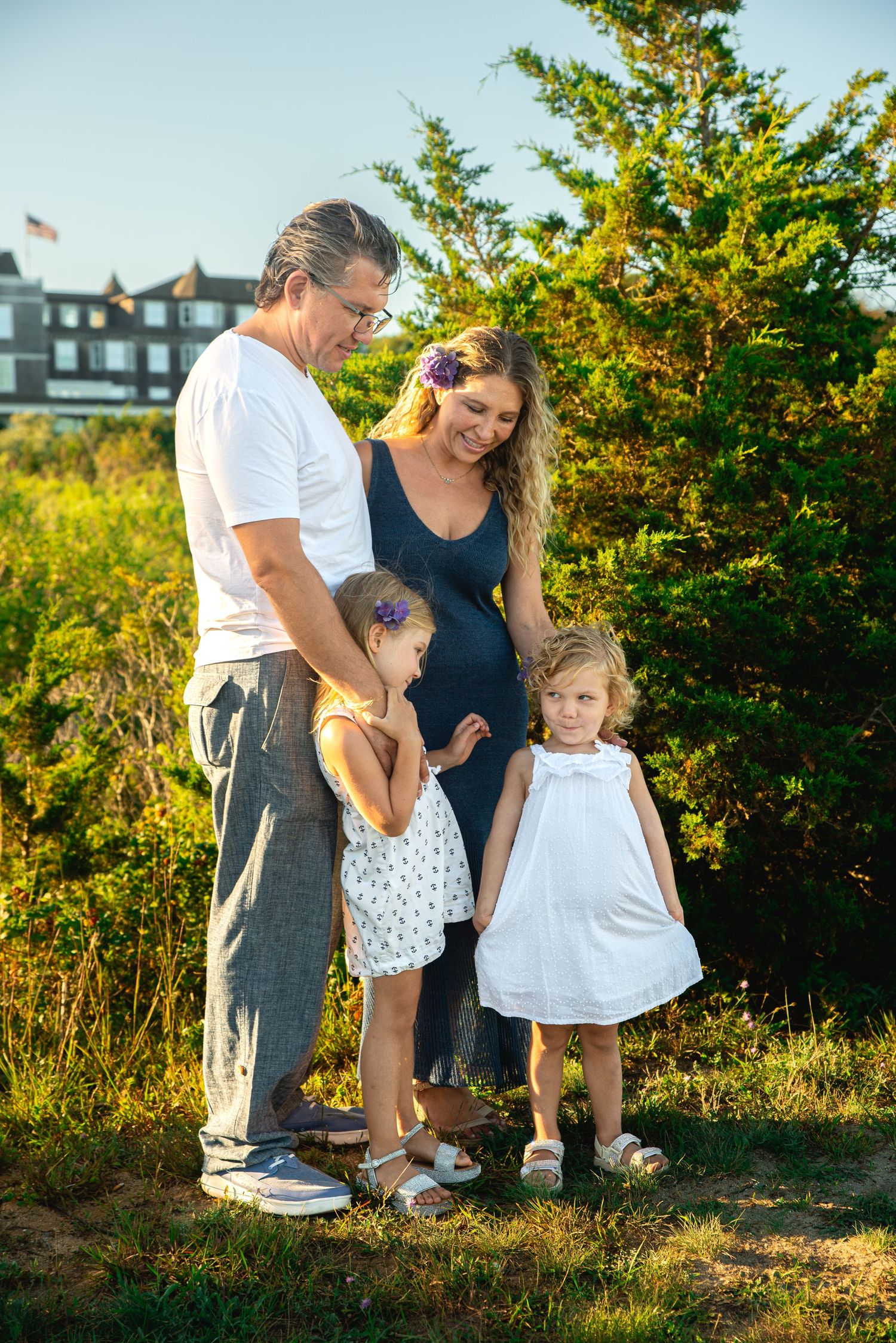 A family of four poses outdoors, with a pregnant mother, father, and two young girls in dresses on a sunny day.