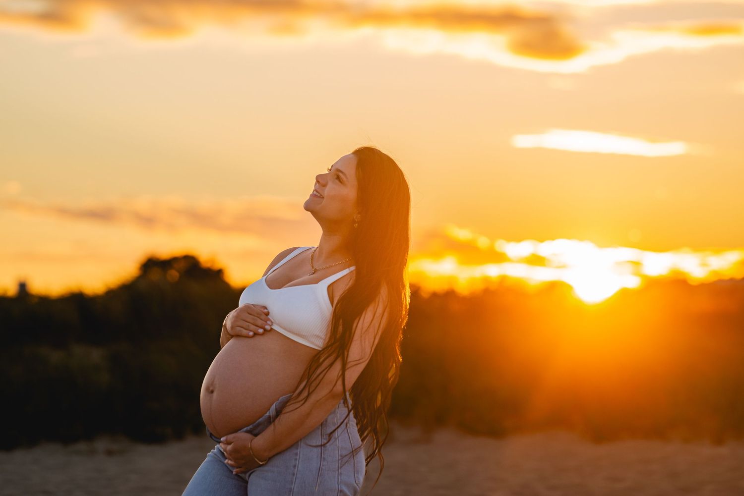 Pregnant woman cradling her baby bump at golden sunset, long dark hair, wearing white crop top and jeans.