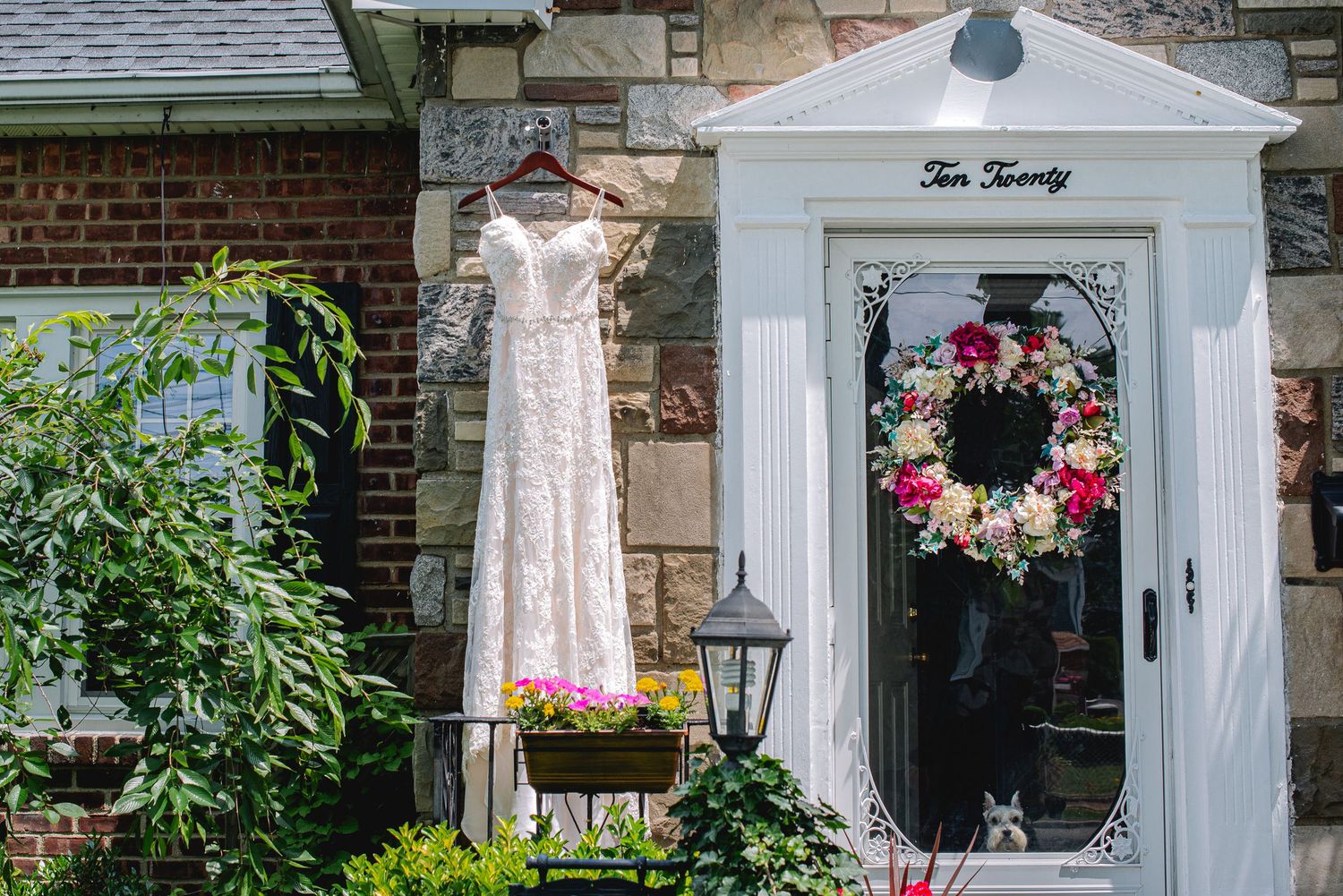 A lace wedding dress hanging outside a brick home with a floral wreath on the white door on a sunny day.
