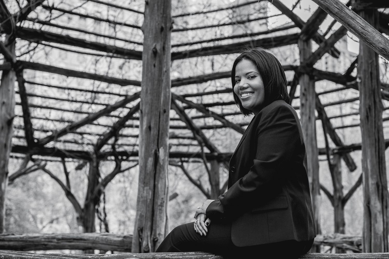 A smiling woman in black sits outdoors under a rustic wooden pergola in a black and white portrait photo.