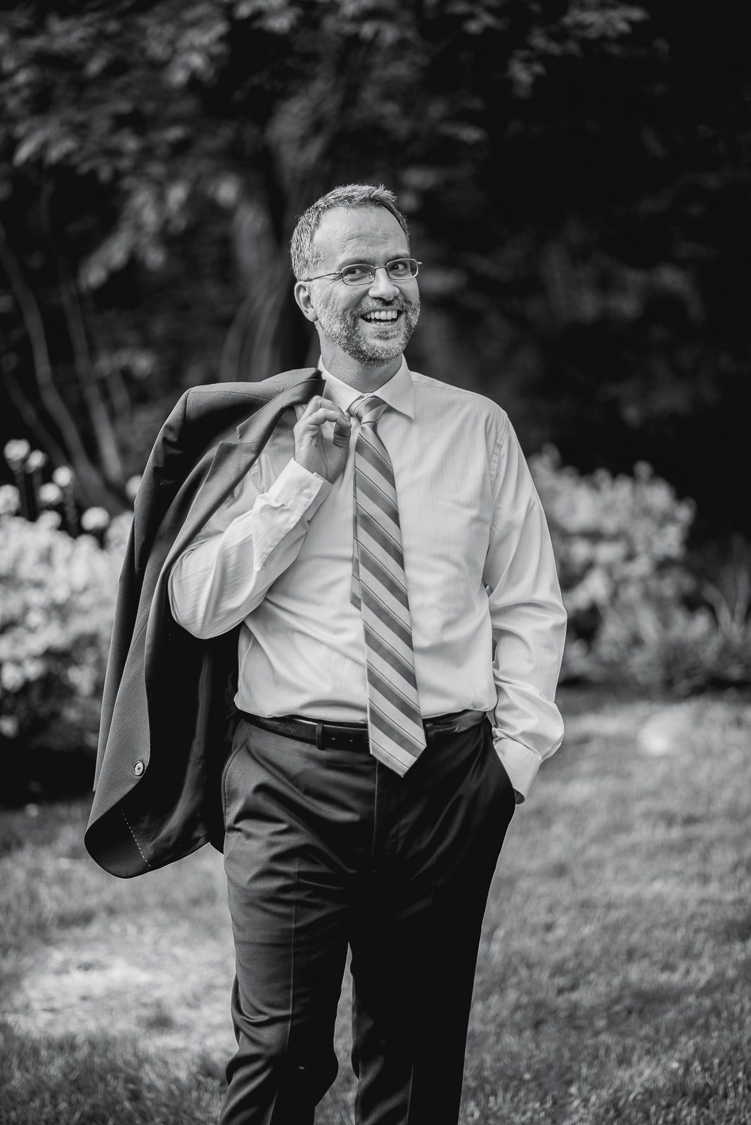 Smiling man in dress shirt and striped tie carries suit jacket over shoulder outdoors in black and white photo.