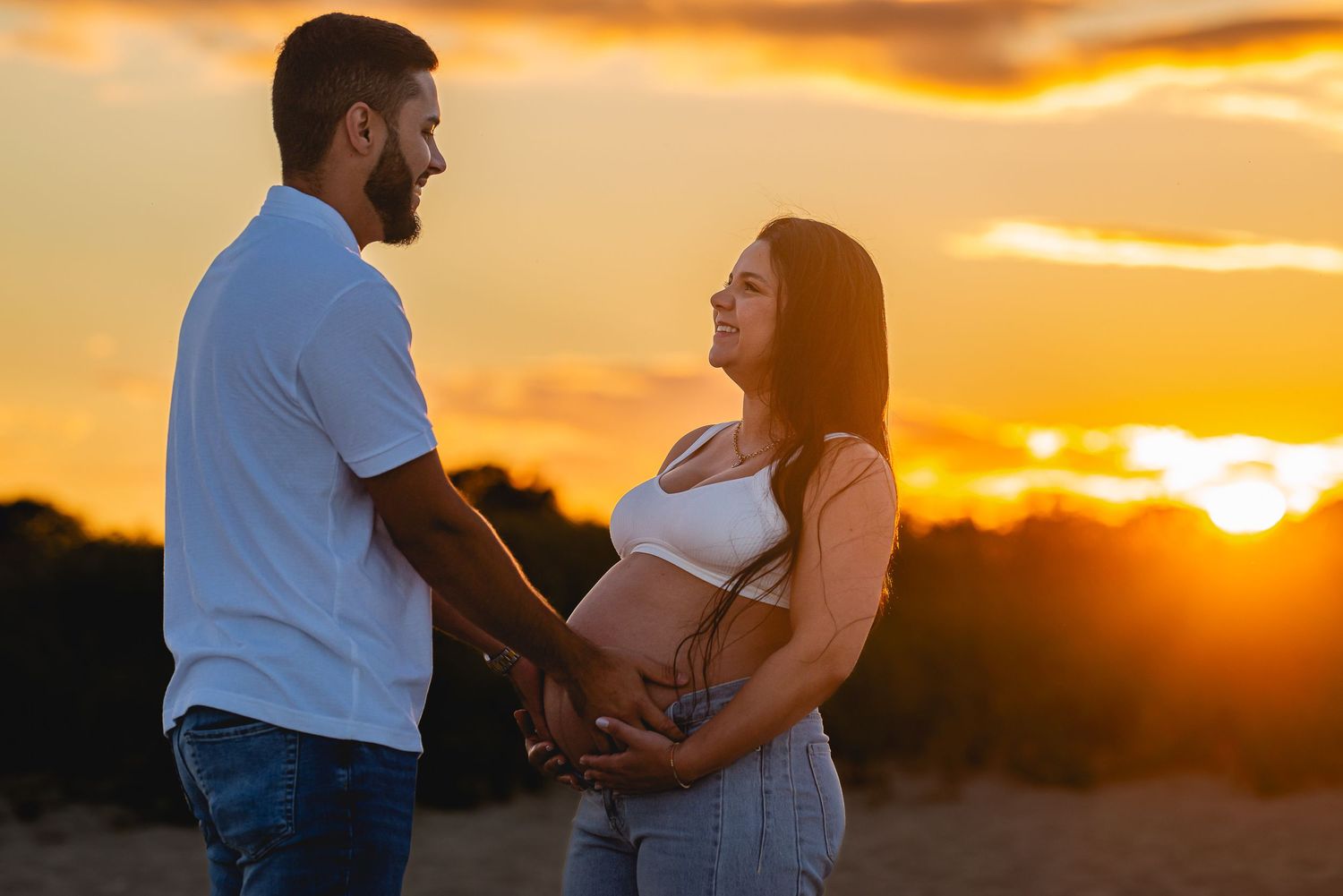 Couple holding pregnant belly at golden sunset in a field, romantic maternity photo outdoors.