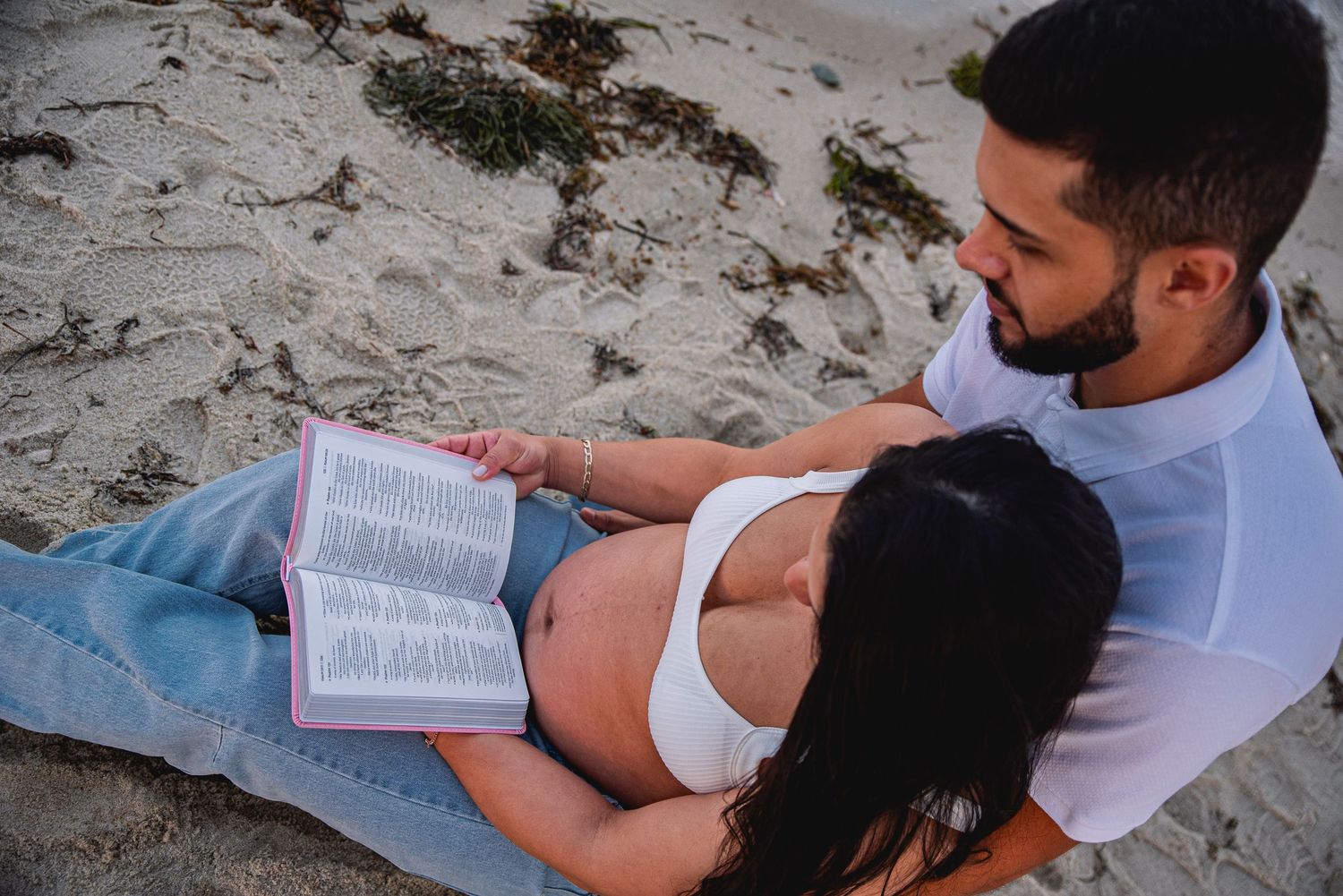 Pregnant couple relaxing on sandy beach, man holding woman as she reads open Bible outdoors.
