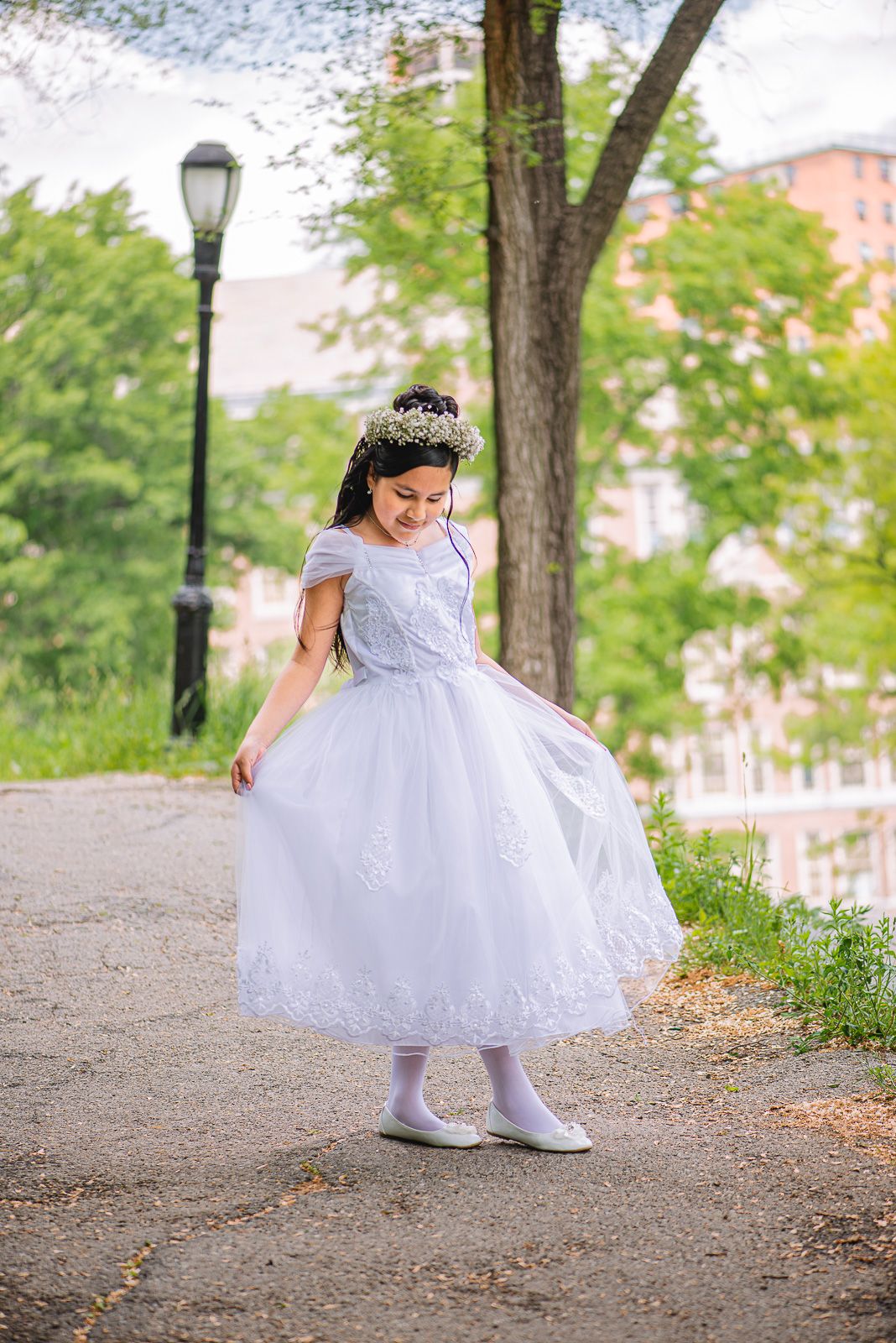 Young girl in white First Communion dress and floral crown twirling on a park path surrounded by green trees.