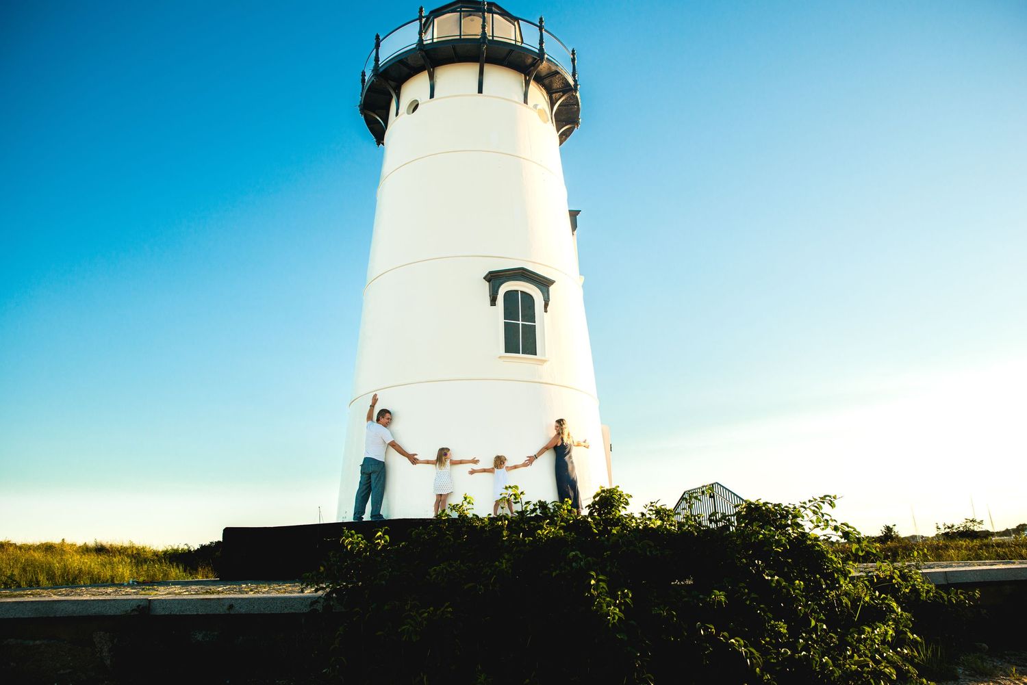 Family holding hands in front of a white lighthouse under a clear blue sky at golden hour.