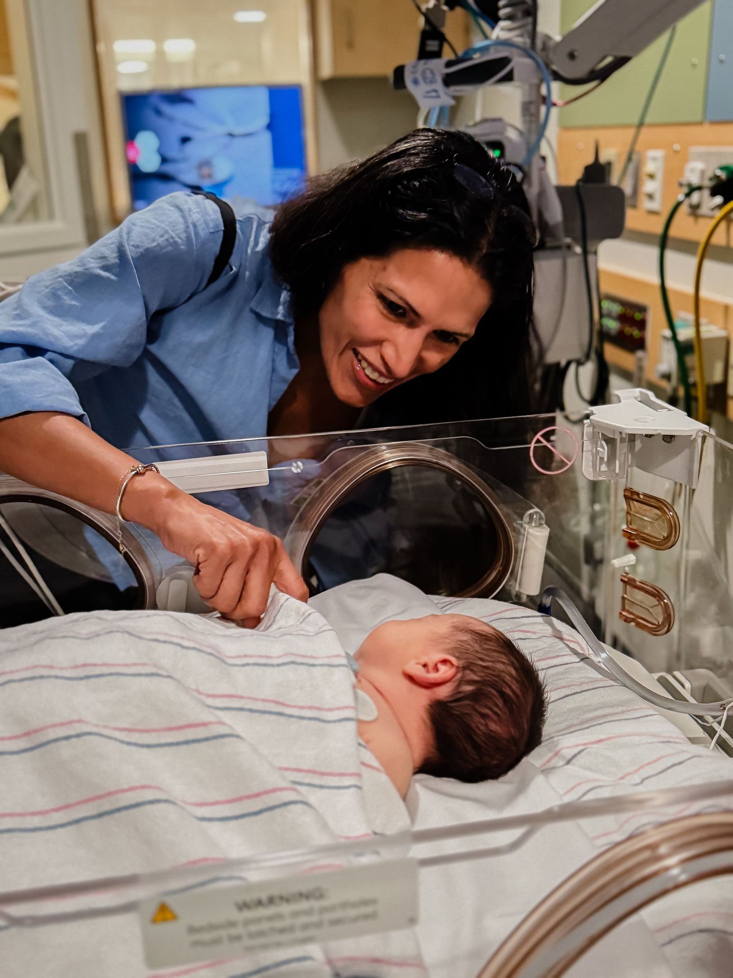 Smiling woman in a blue top leans over a sleeping newborn baby wrapped in a striped blanket inside a hospital incubator in a neonatal unit.