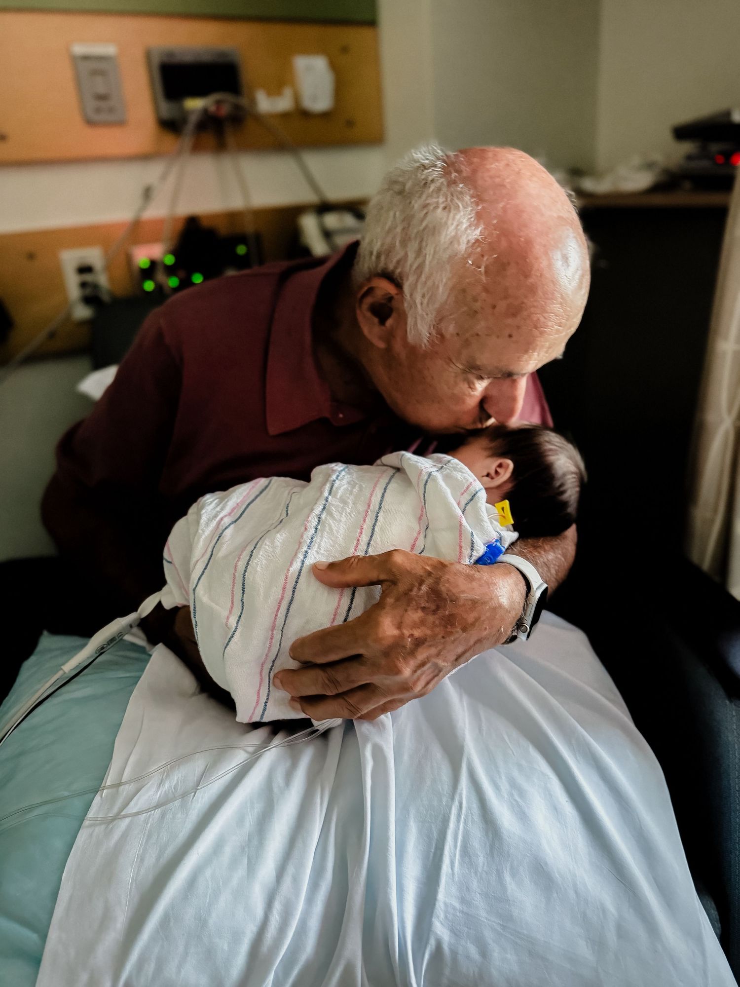 Elderly man in a maroon shirt gently kisses a newborn baby wrapped in a hospital blanket while holding the infant in a hospital room.