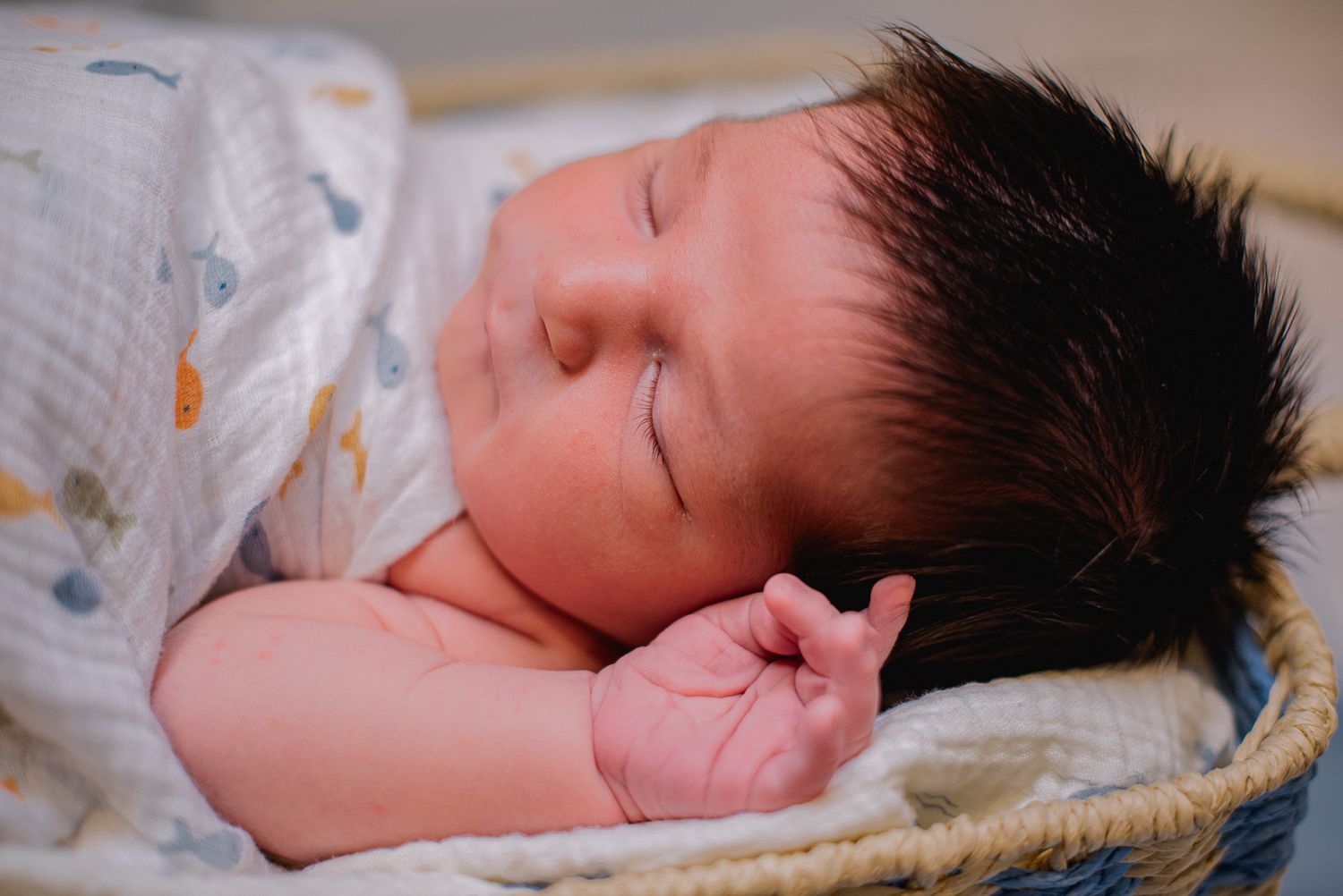 Peaceful newborn baby with dark hair sleeping in a wicker basket, wrapped in a soft white swaddle blanket.