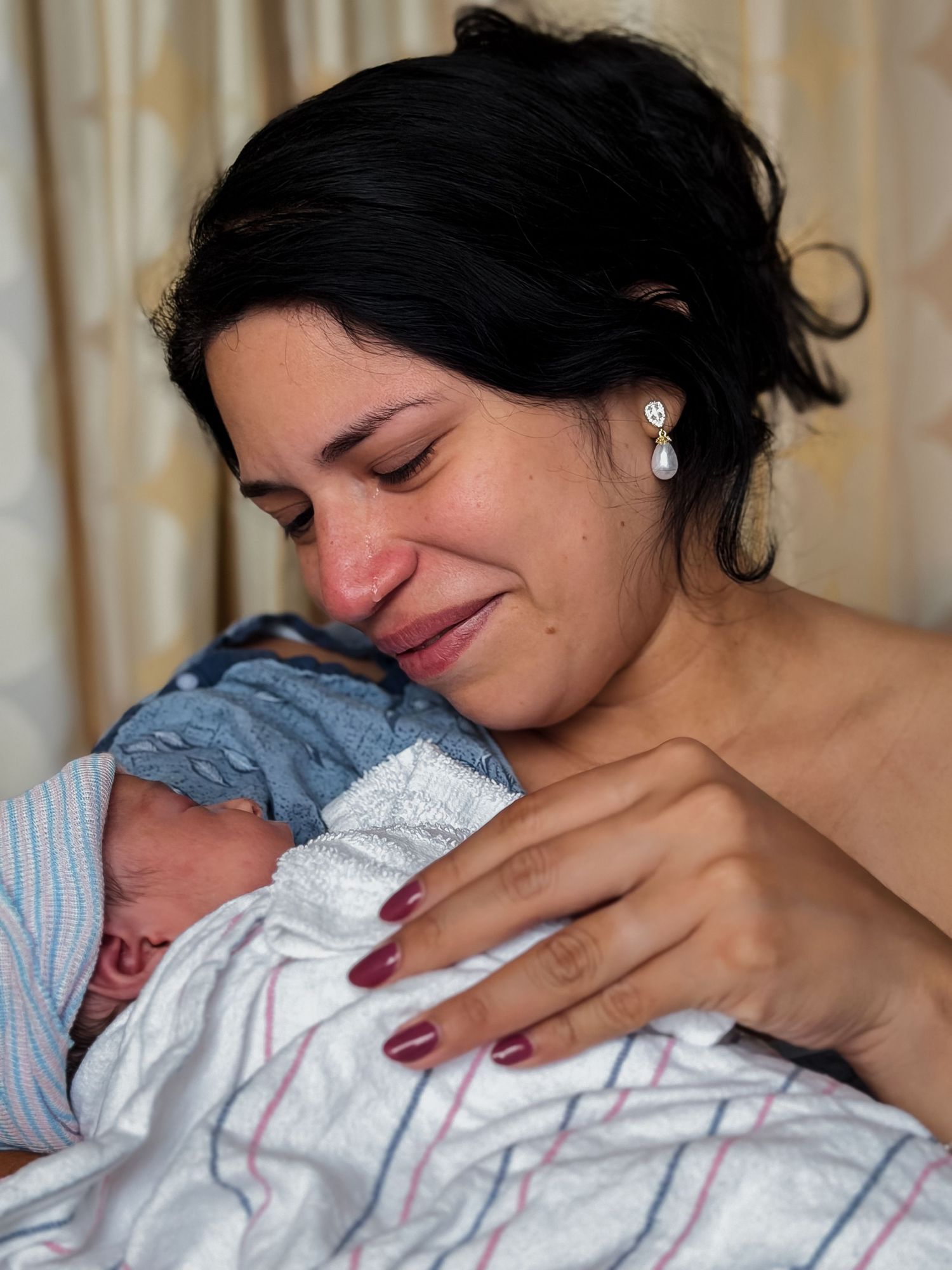 Emotional new mother with dark hair and pearl earrings smiles tearfully while cradling her newborn baby wrapped in a hospital blanket.