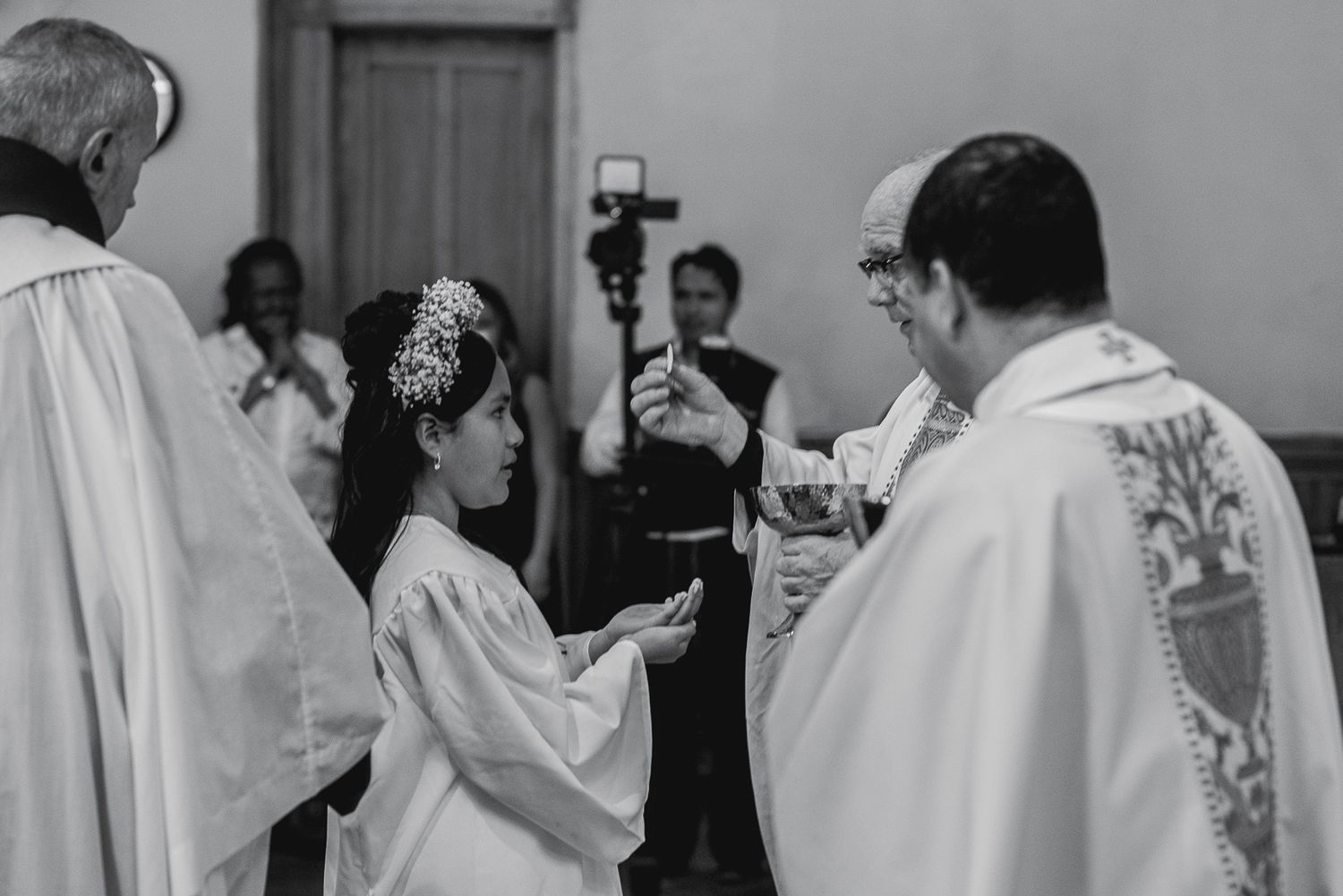Black and white photo of a child in white dress receiving First Communion from a priest in a church ceremony.