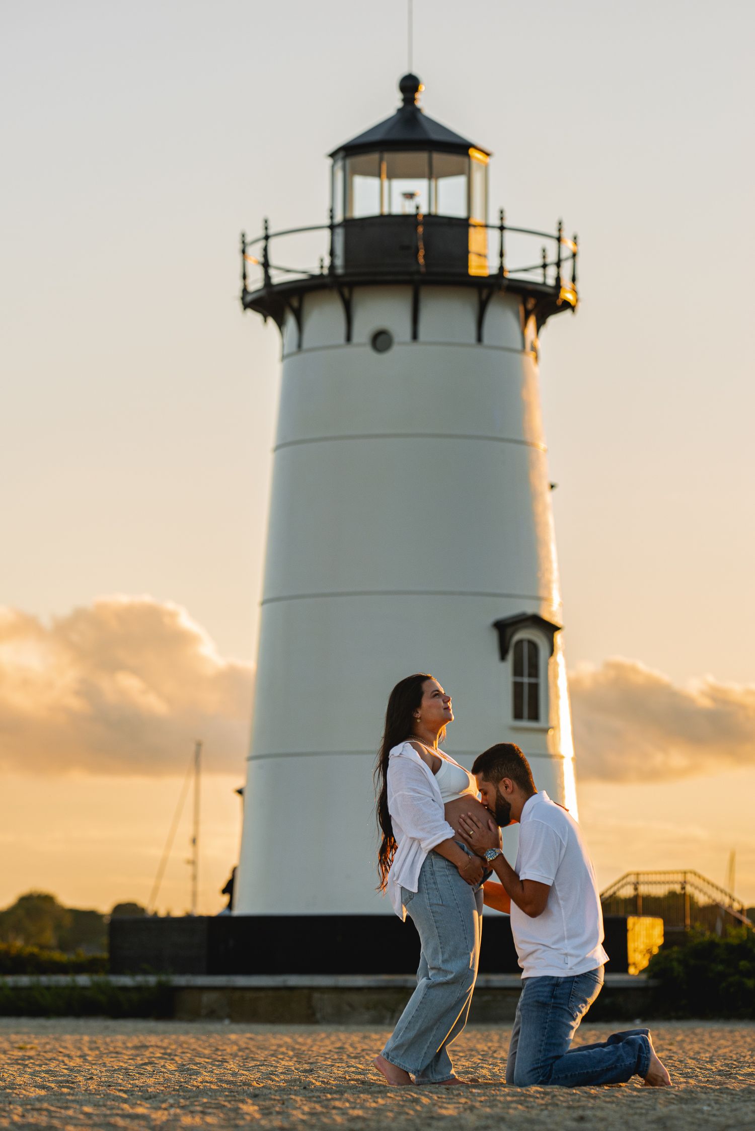 A man kneeling before a woman at sunset near a white lighthouse, appearing to propose on a sandy beach.