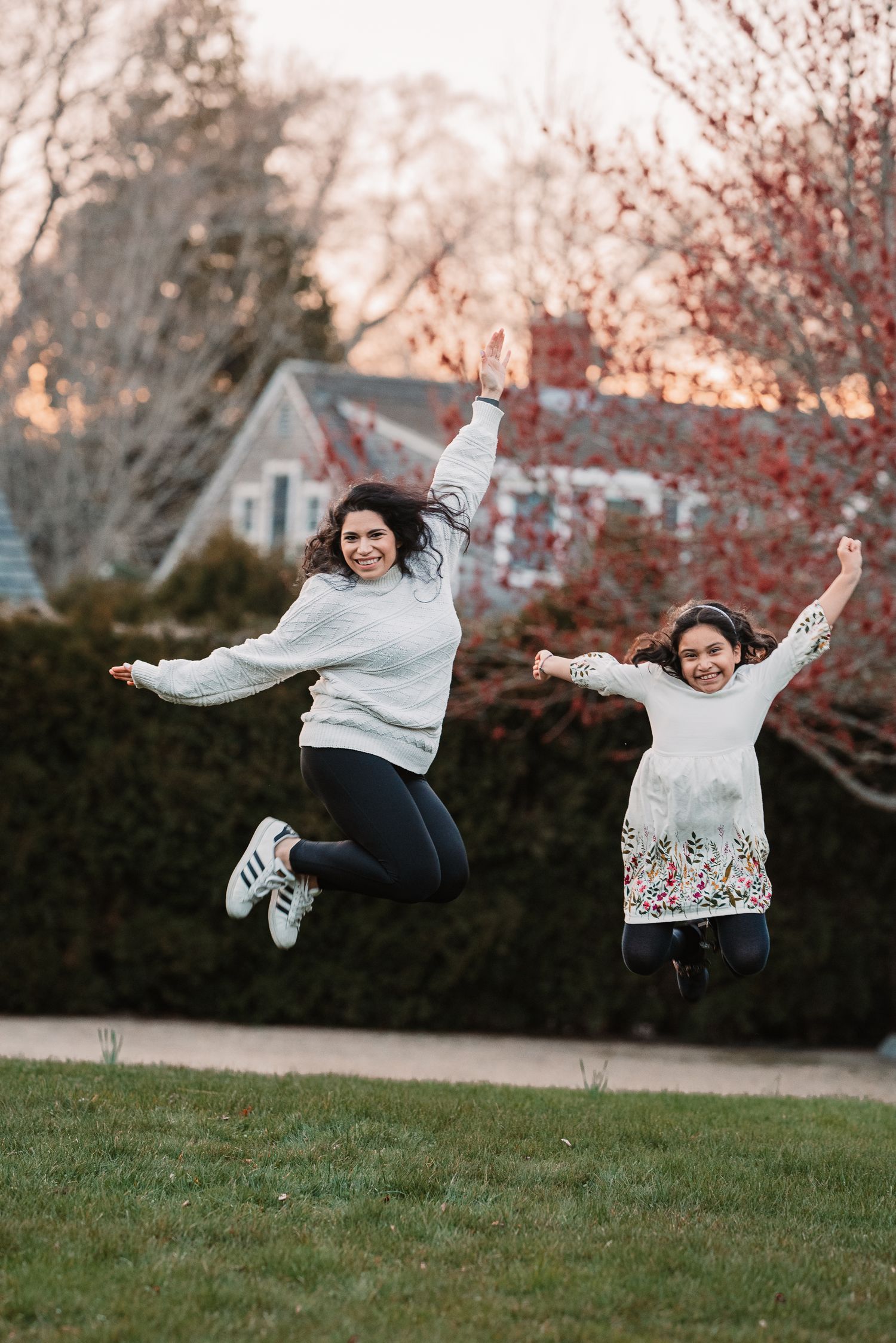 Mother and daughter jumping joyfully in yard at sunset with arms raised.