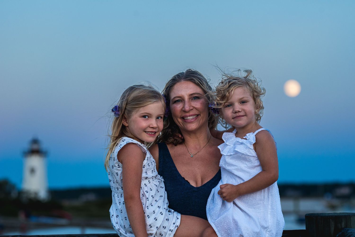 Woman smiling with two young girls near a lighthouse at dusk with a full moon rising in the background.