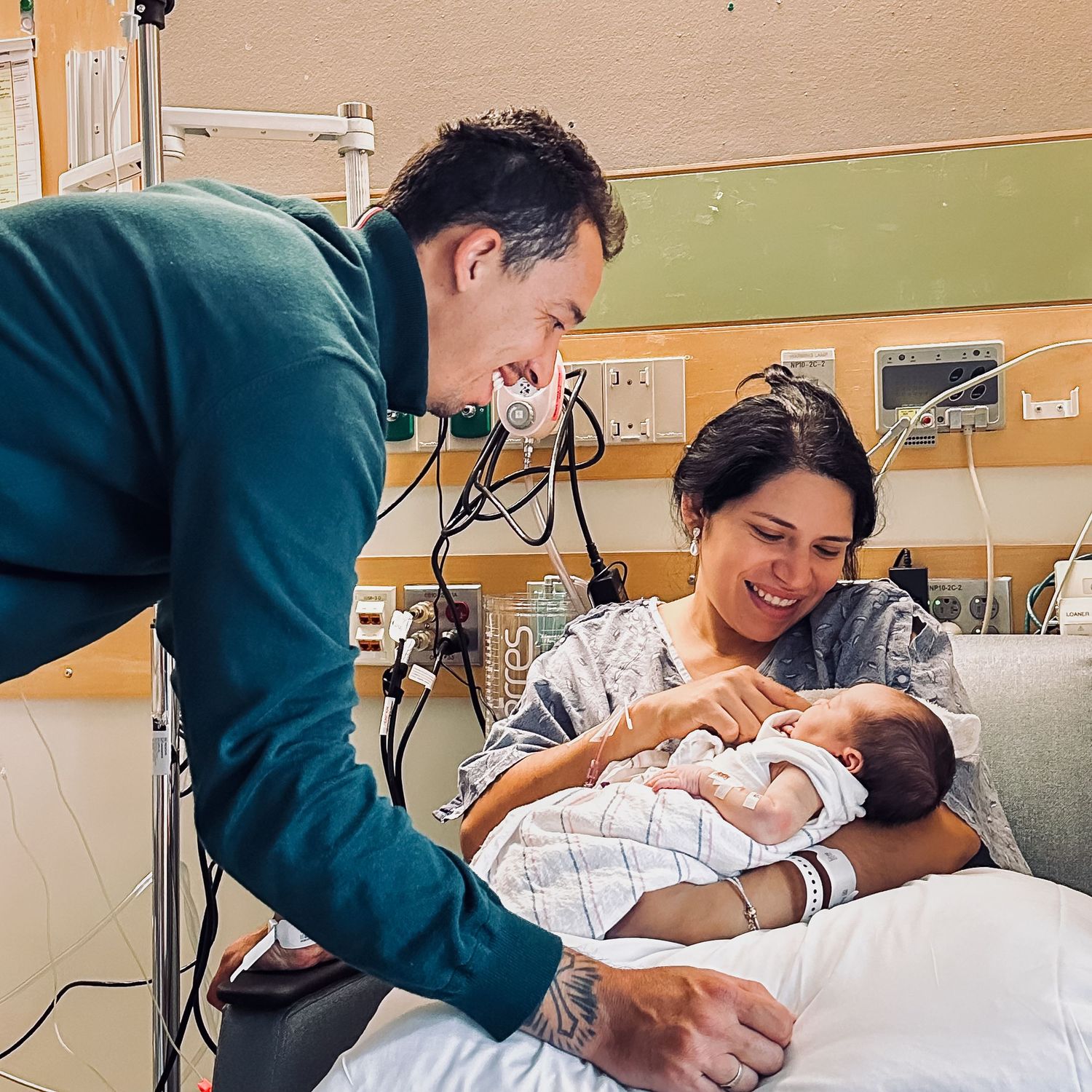 Smiling new parents in a hospital room, with a mother in a gown cradling her newborn baby while the father leans over to look on.