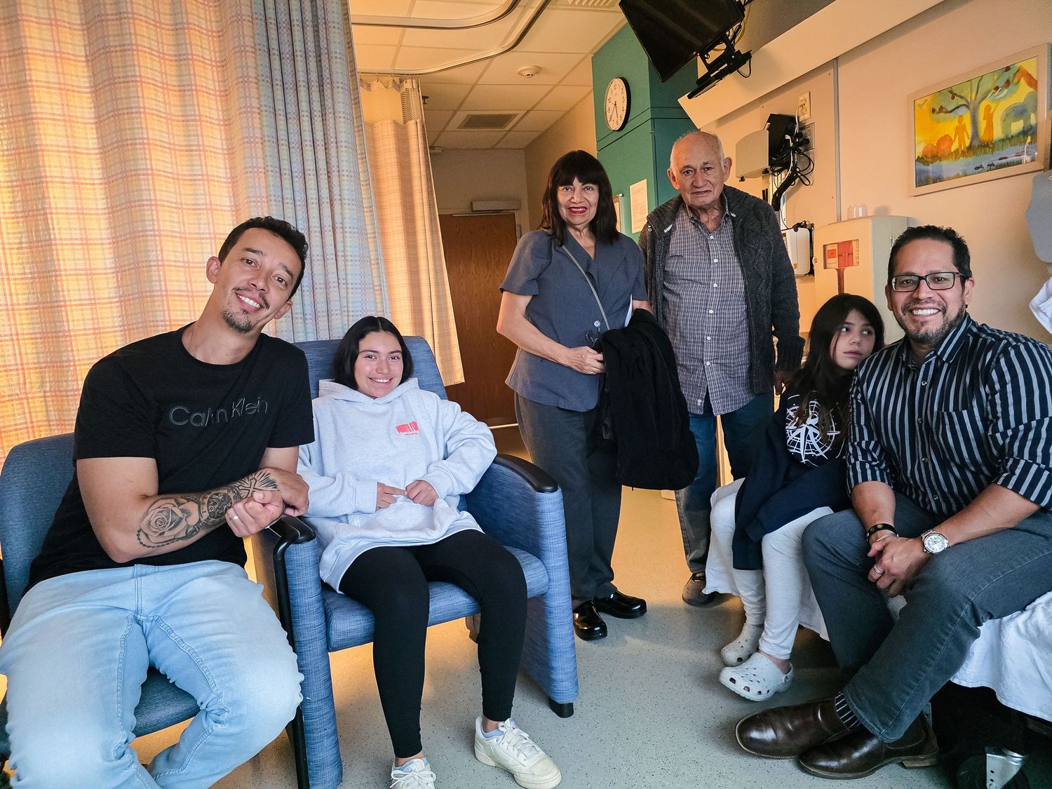 Six people, including a young girl in a white hoodie seated in a blue chair, gather and smile together in a bright hospital room.