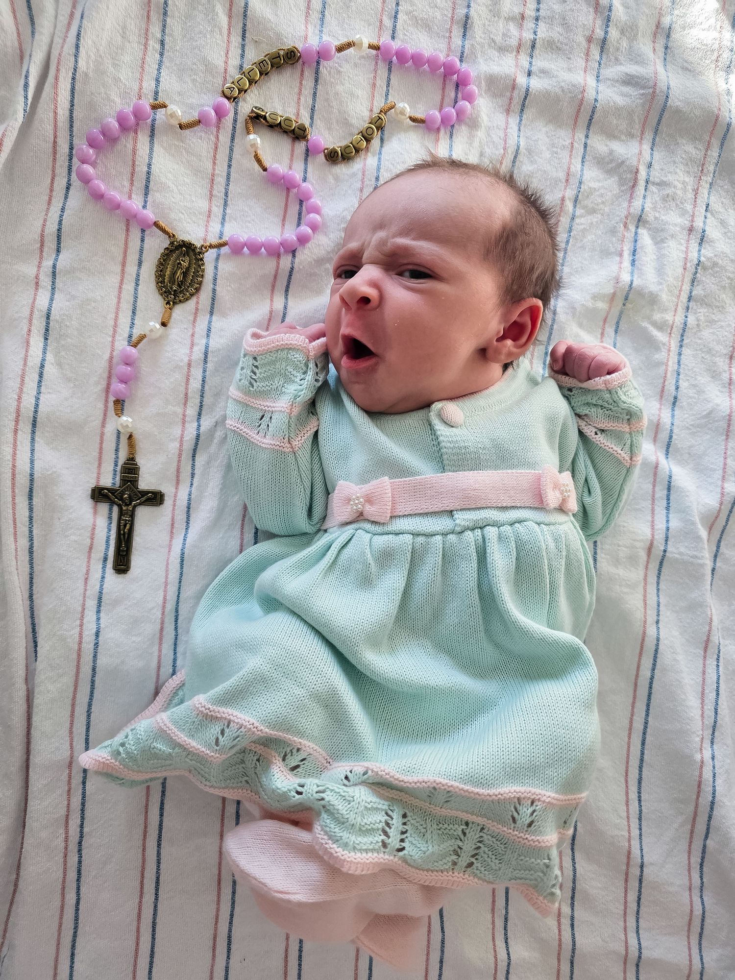 Newborn baby in a mint green knit dress yawning on a striped blanket beside a pink and gold rosary with a crucifix.