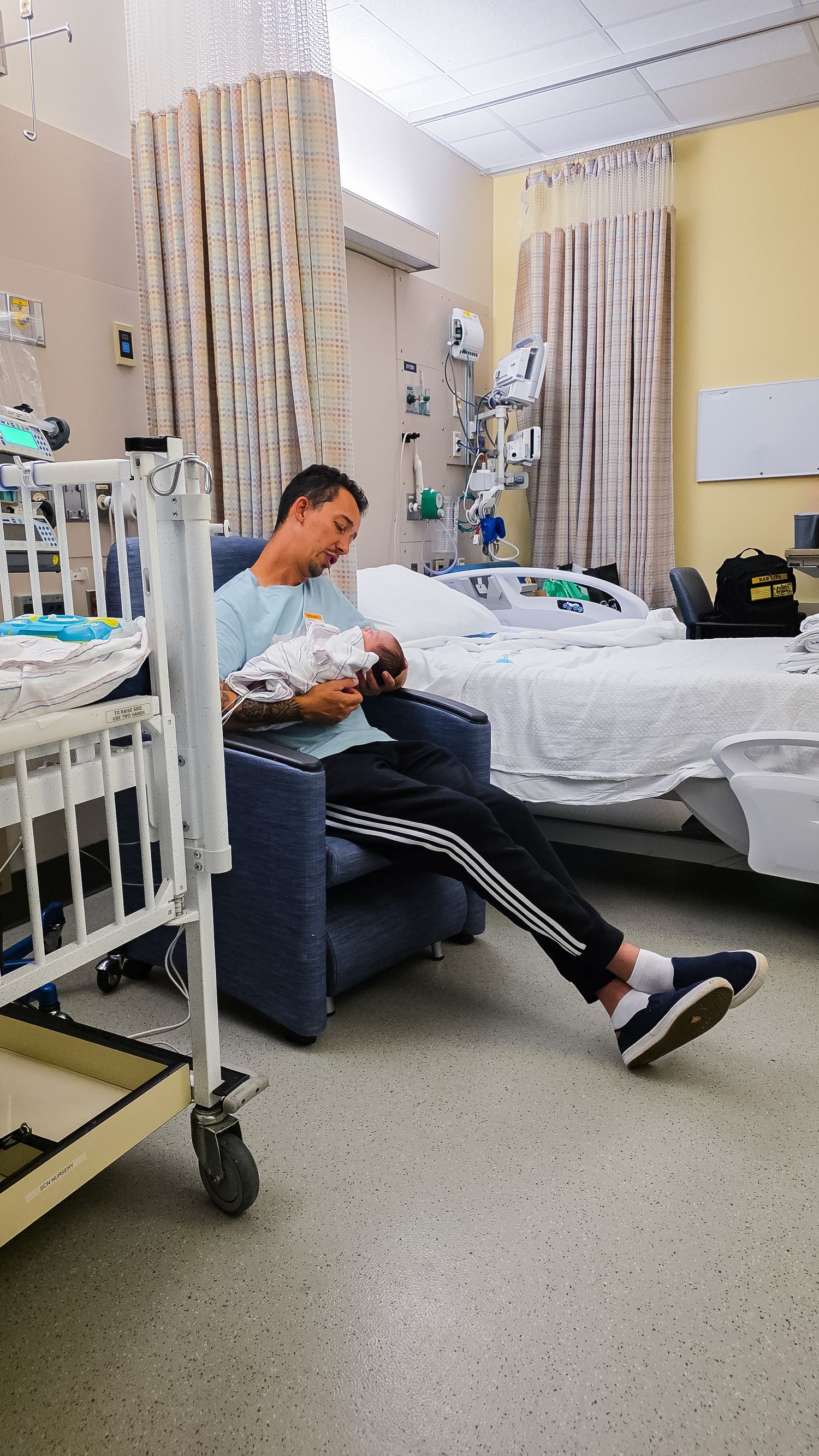 A father in a blue shirt and track pants sits in a recliner in a hospital room, cradling his newborn baby wrapped in a blanket.