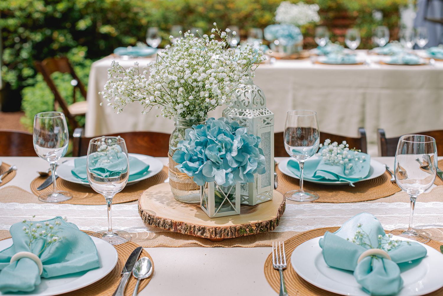 Elegant outdoor table setting with teal napkins, burlap chargers, crystal glasses, and floral centerpiece.