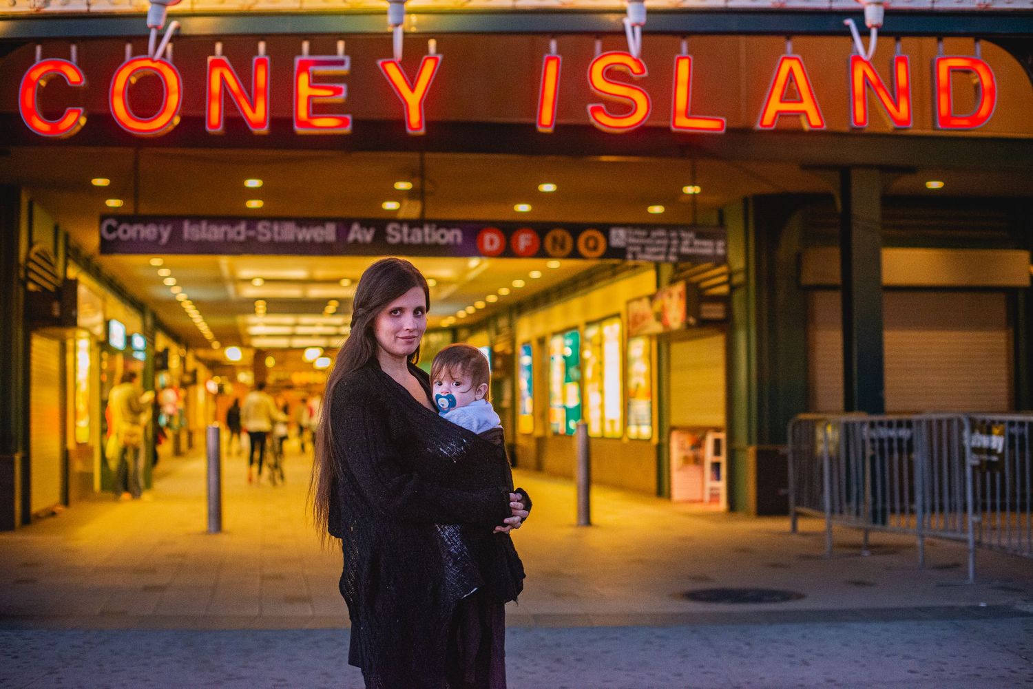 Woman holding baby outside Coney Island-Stillwell Ave Station at night with bright neon signs.