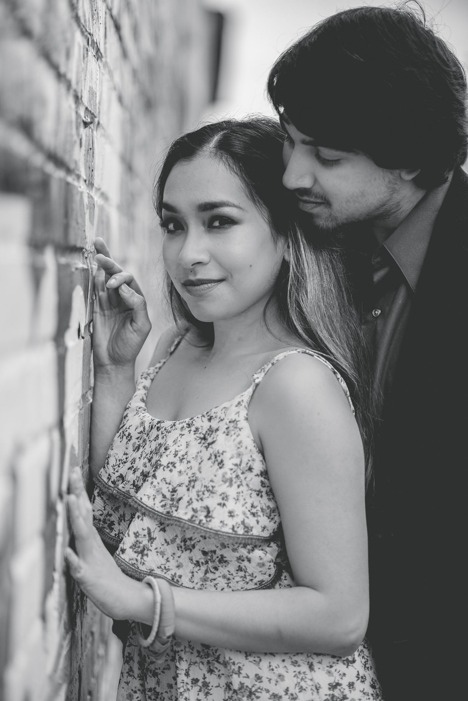 Black and white portrait of a couple leaning against a brick wall, woman in floral dress smiling softly.