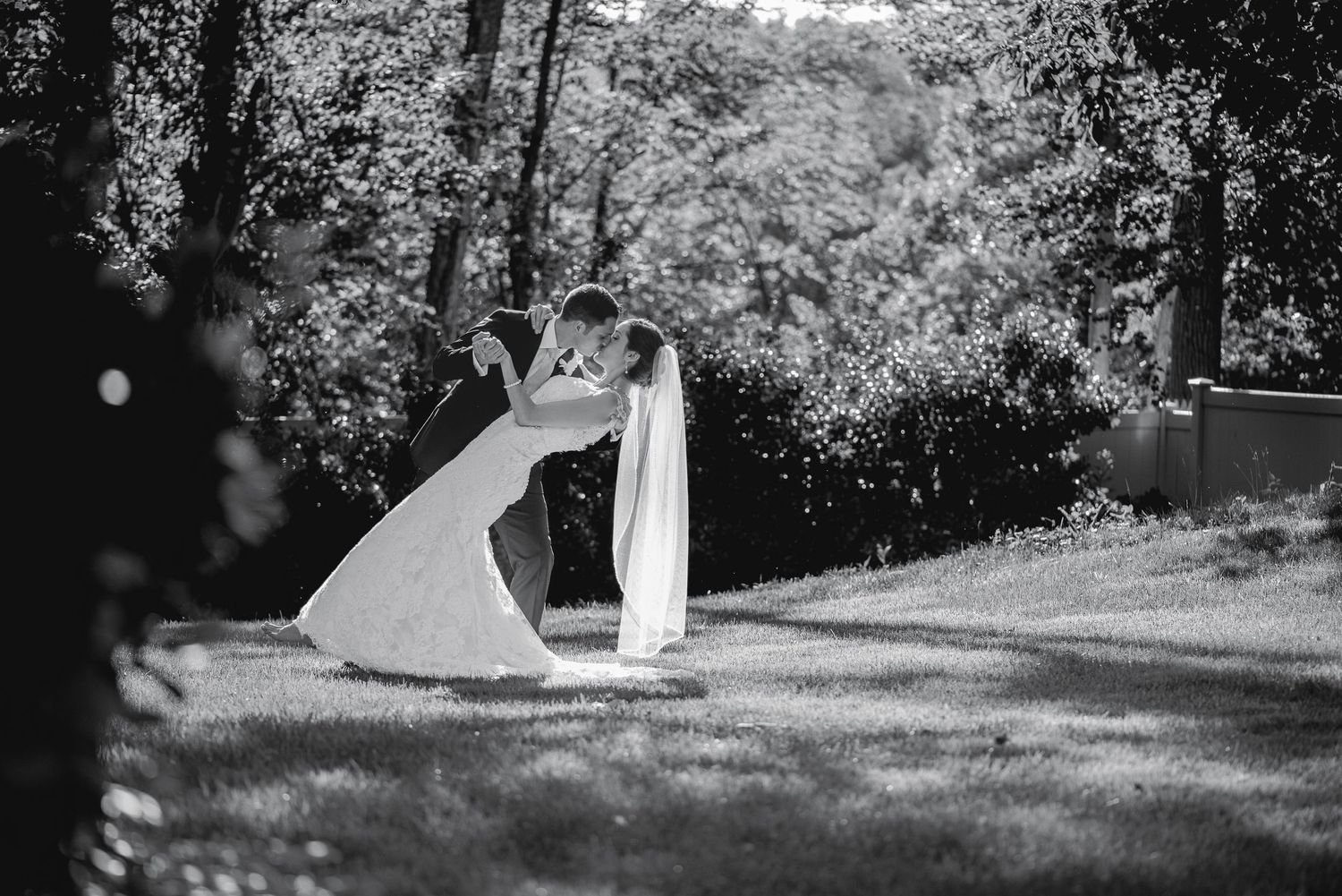 Black and white photo of a bride and groom sharing a romantic dip kiss outdoors surrounded by lush trees.