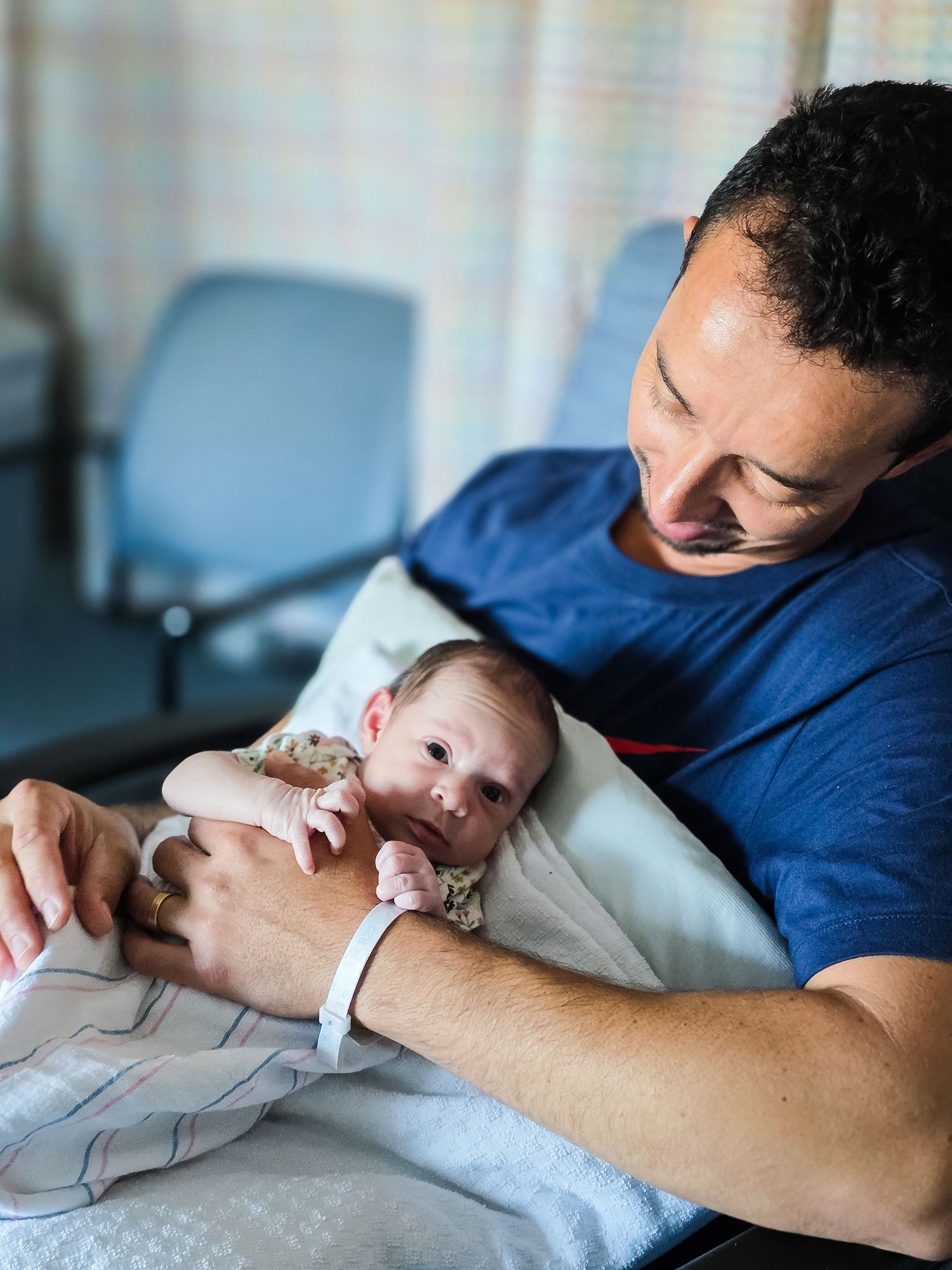Smiling father in a blue t-shirt gently cradles his wide-eyed newborn baby on a pillow in a hospital room.