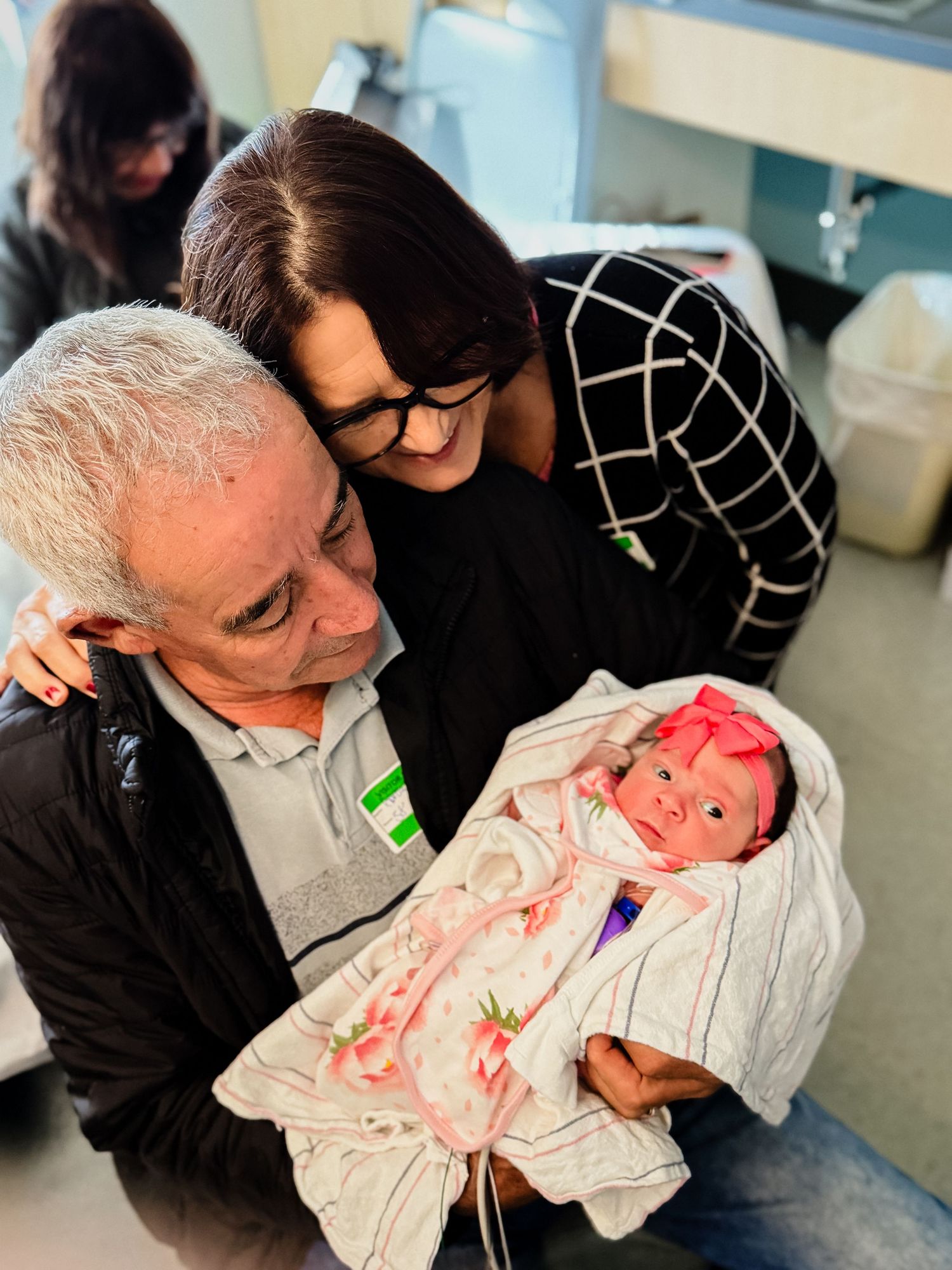 Smiling grandparents in a hospital room lovingly hold a newborn baby wrapped in a pink floral blanket with a pink bow headband.