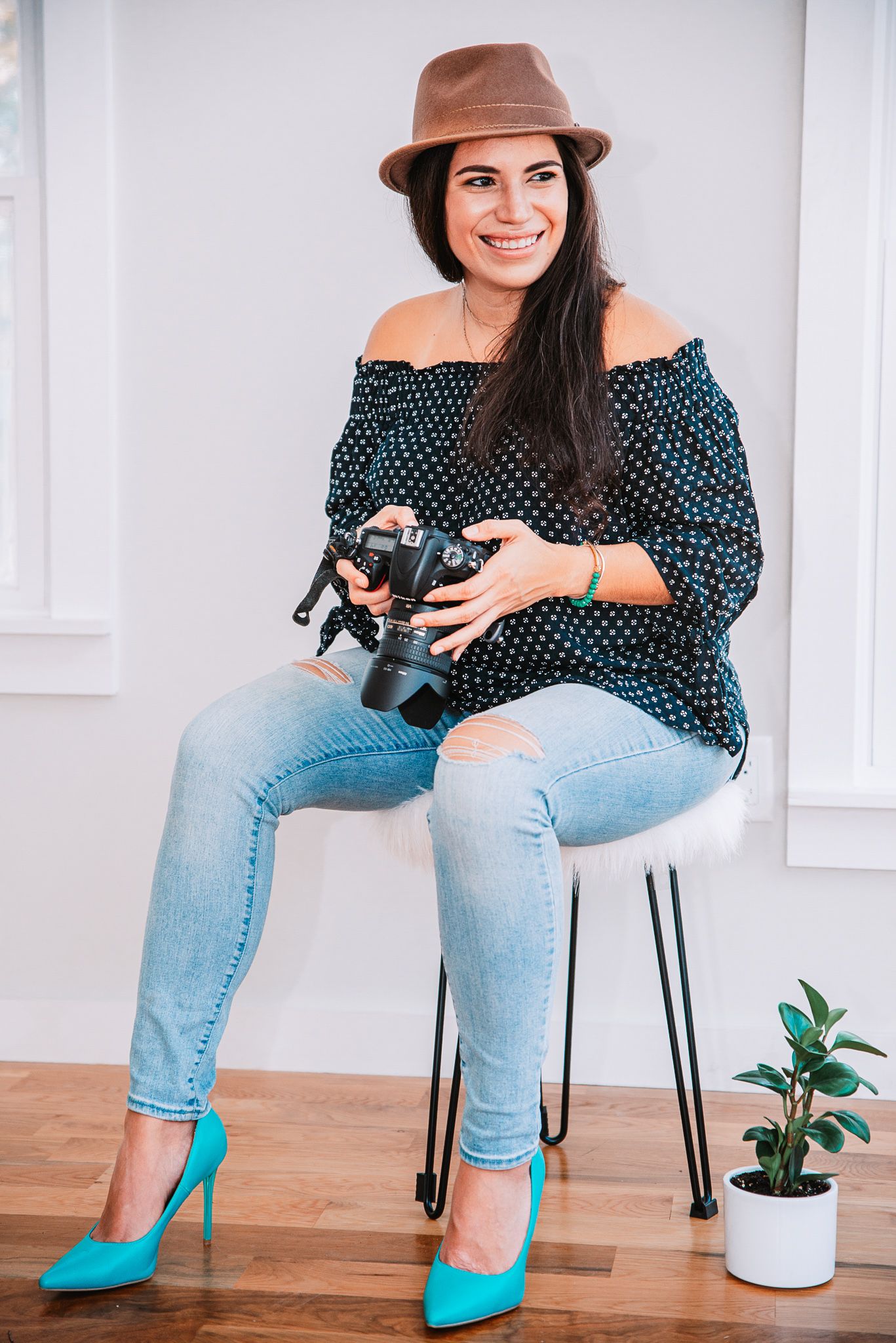 Smiling female photographer in hat and teal heels sitting with DSLR camera.