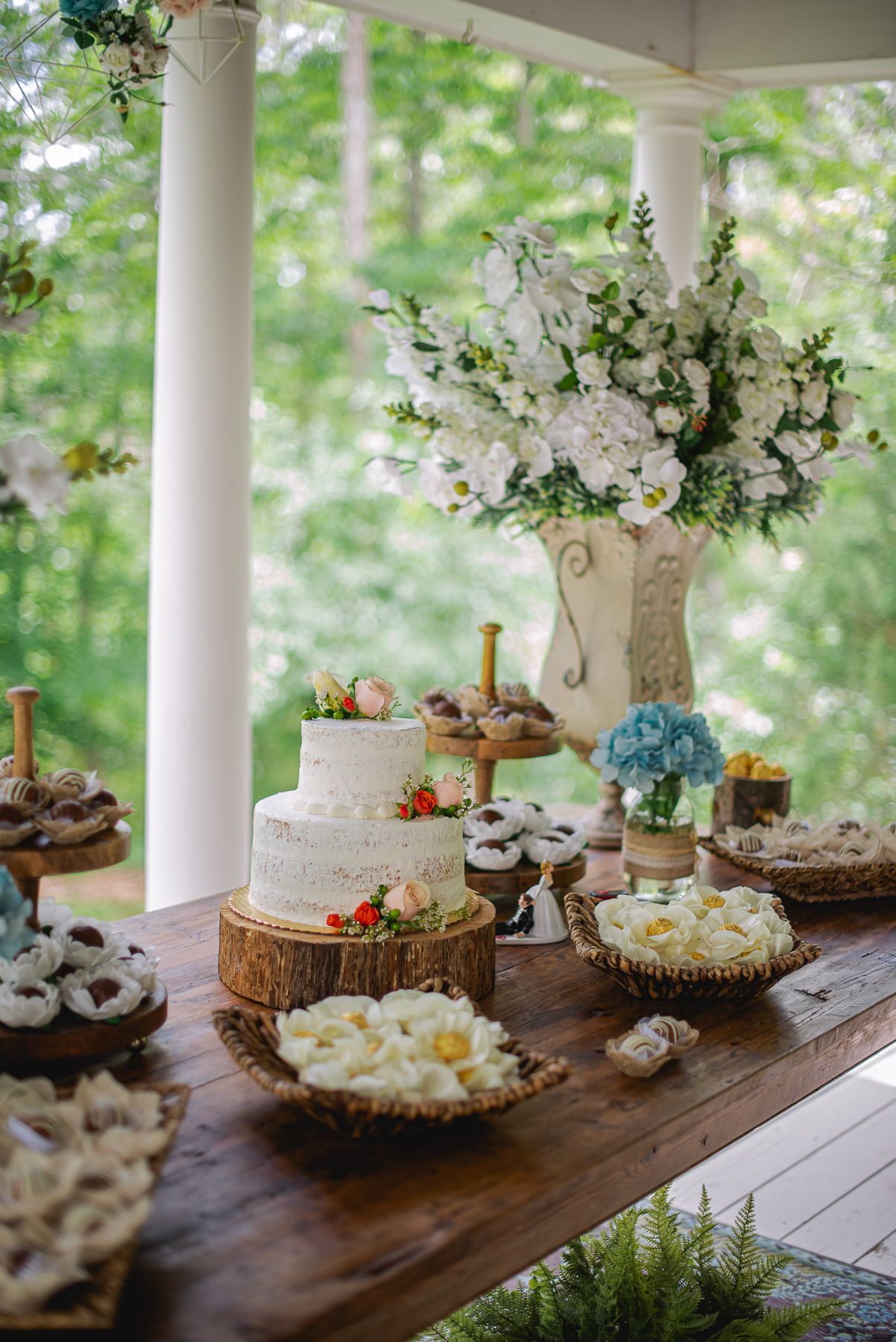 Rustic wedding dessert table with white tiered cake on wood slice, floral arrangements, and assorted sweets on wooden table.