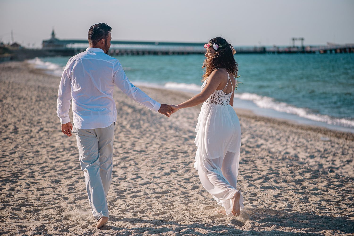 Bride and groom in white walking hand in hand along a sandy beach near a pier at sunset.