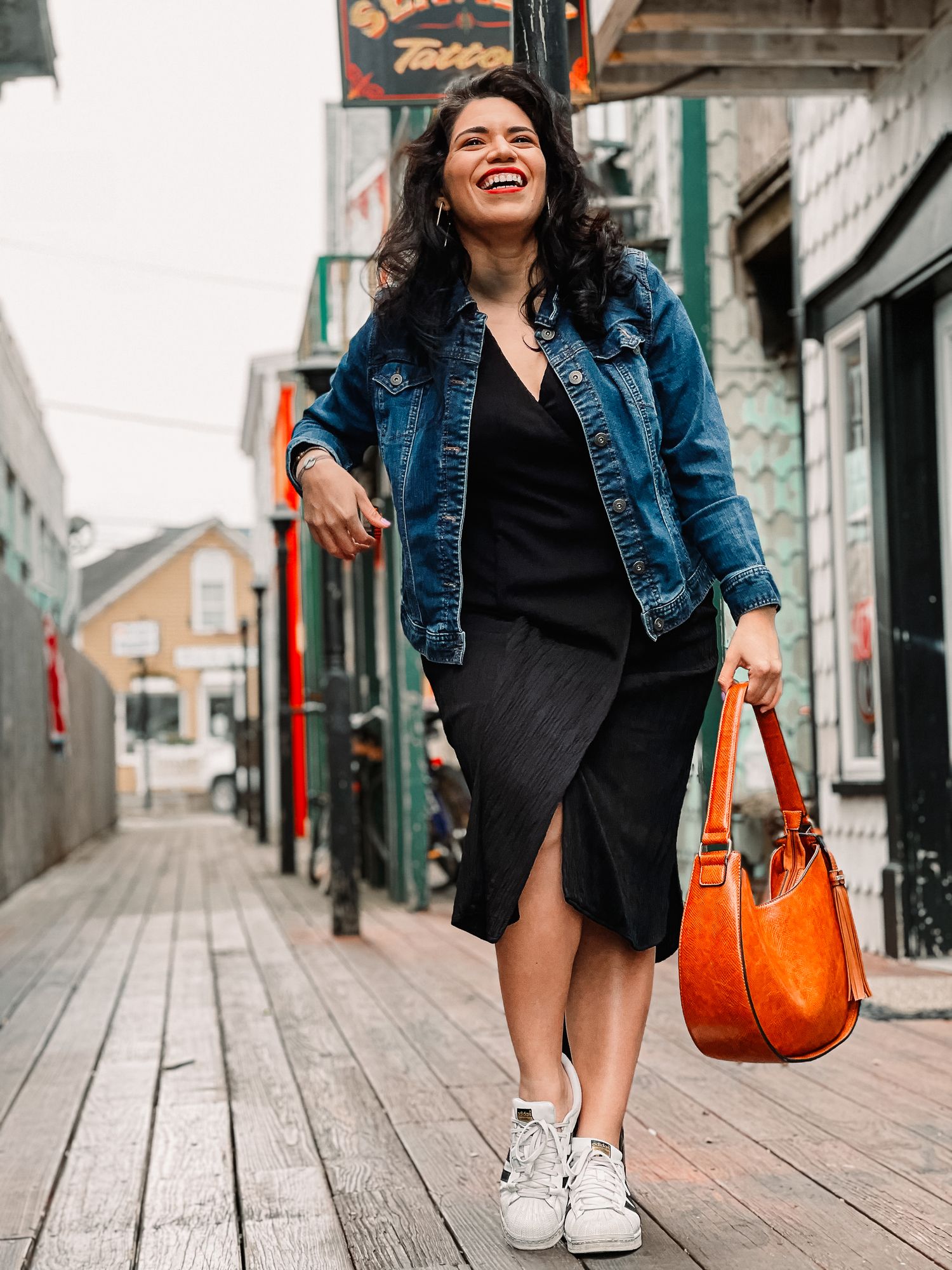 Happy curly-haired woman in denim jacket and black dress walking on a wooden boardwalk with orange bag.