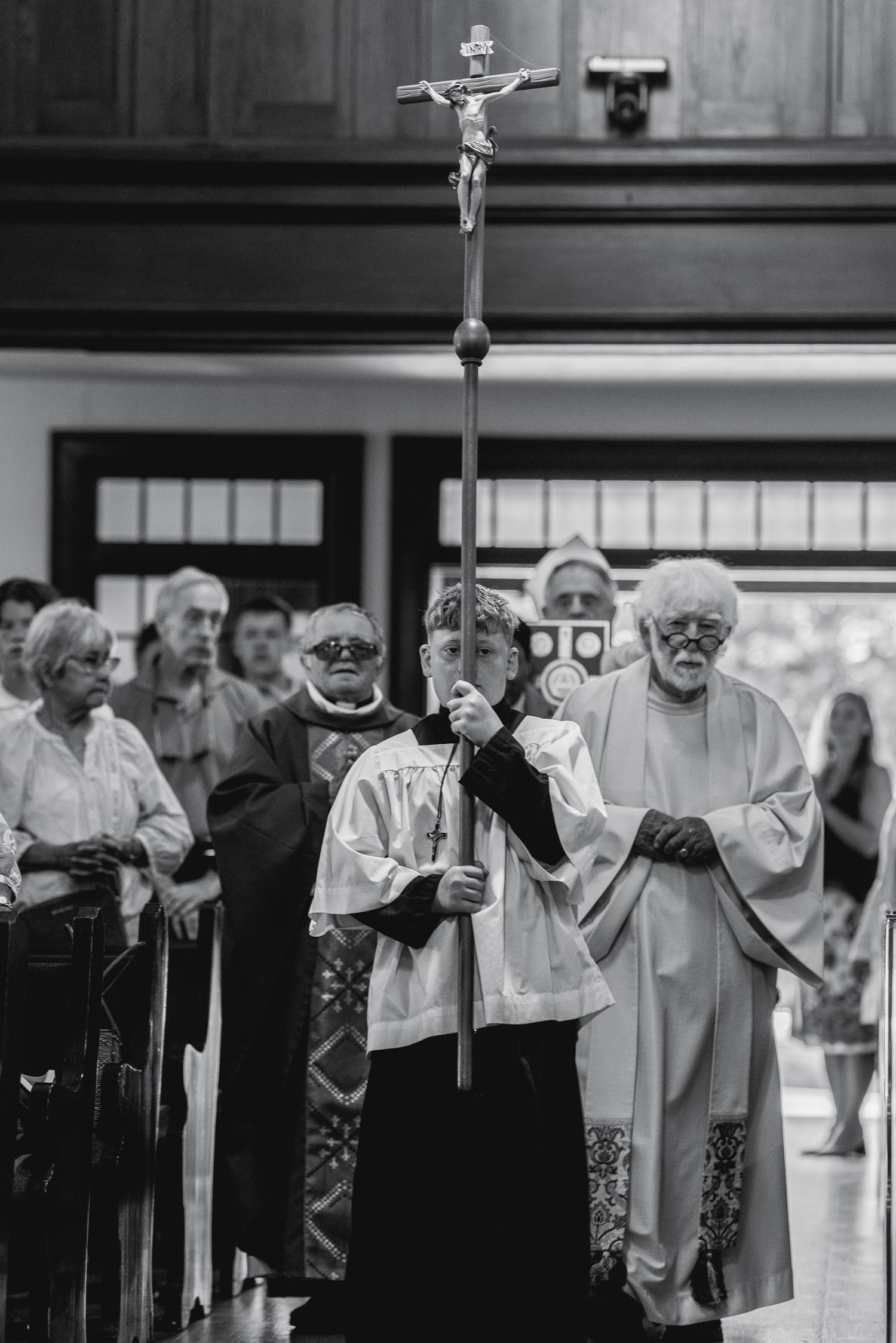 Black and white photo of Catholic priests leading a processional with a crucifix staff inside a church.