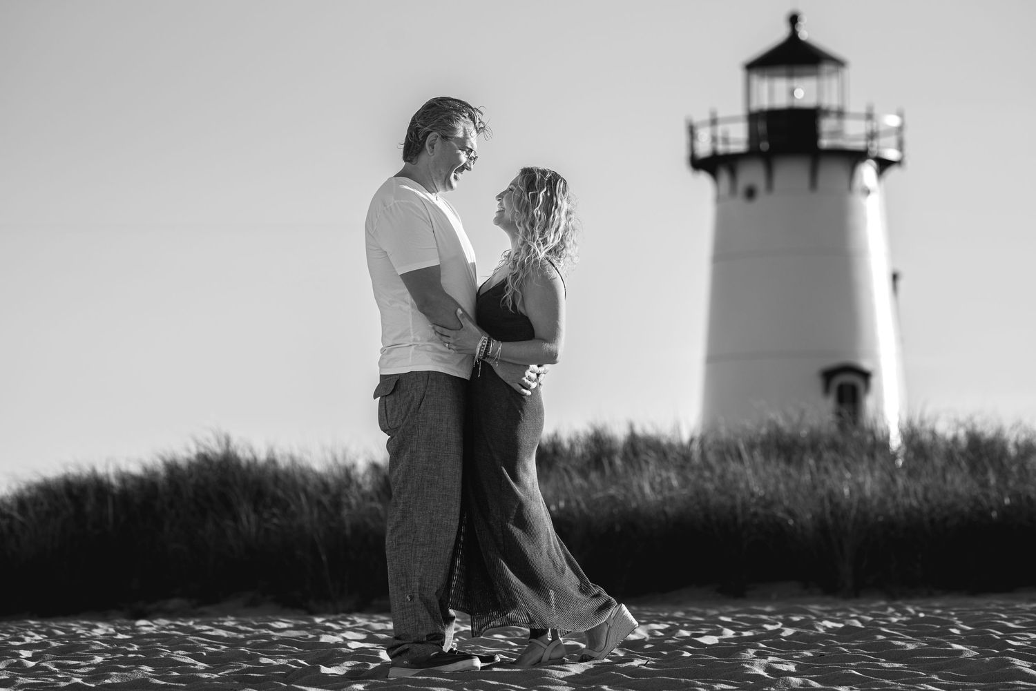 Couple embracing near a lighthouse in a black and white beach photo.