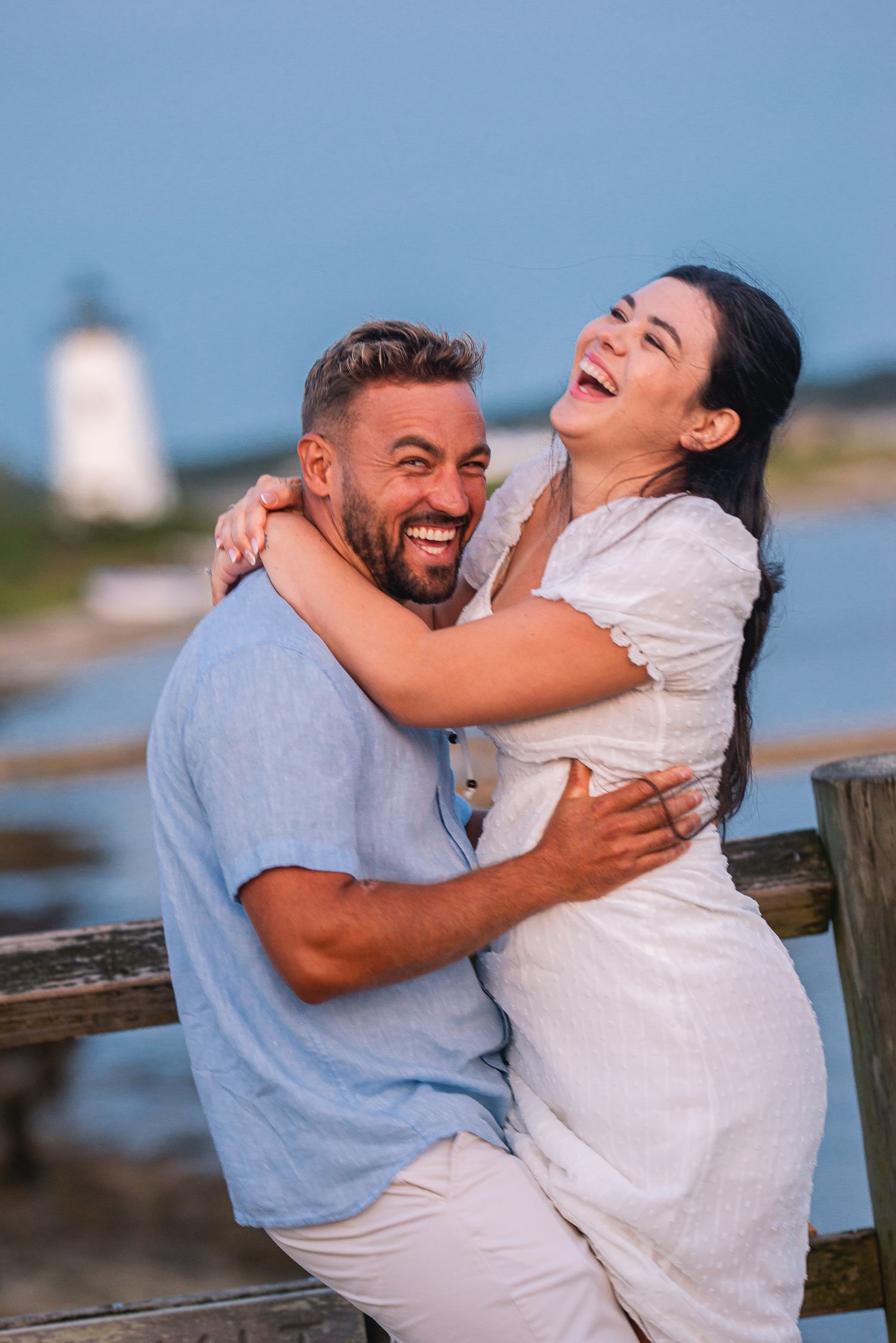 Happy couple laughing and embracing on a wooden dock near a lighthouse by the water on a sunny day.