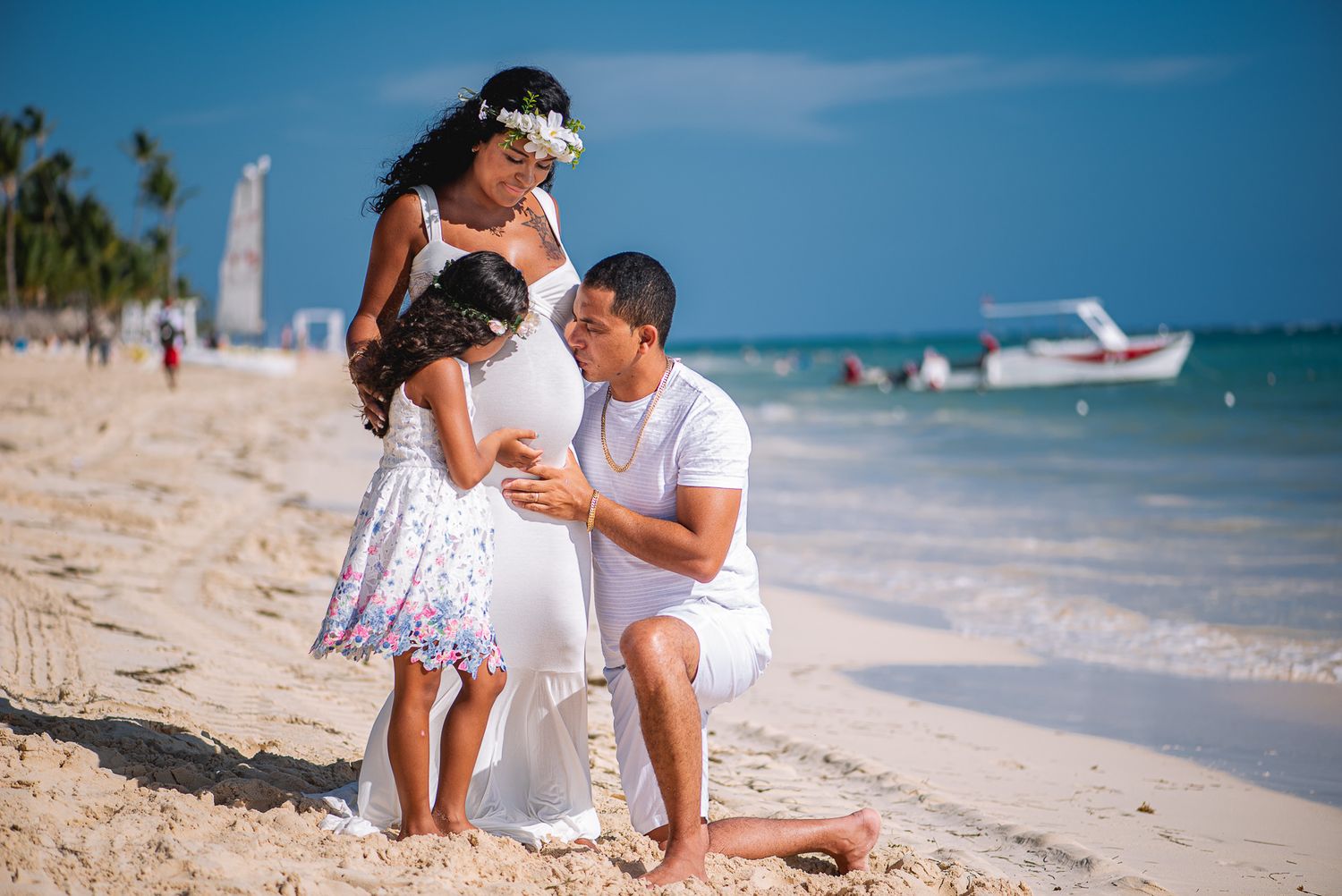 Family maternity photo on tropical beach with pregnant woman, man, and young girl in white.