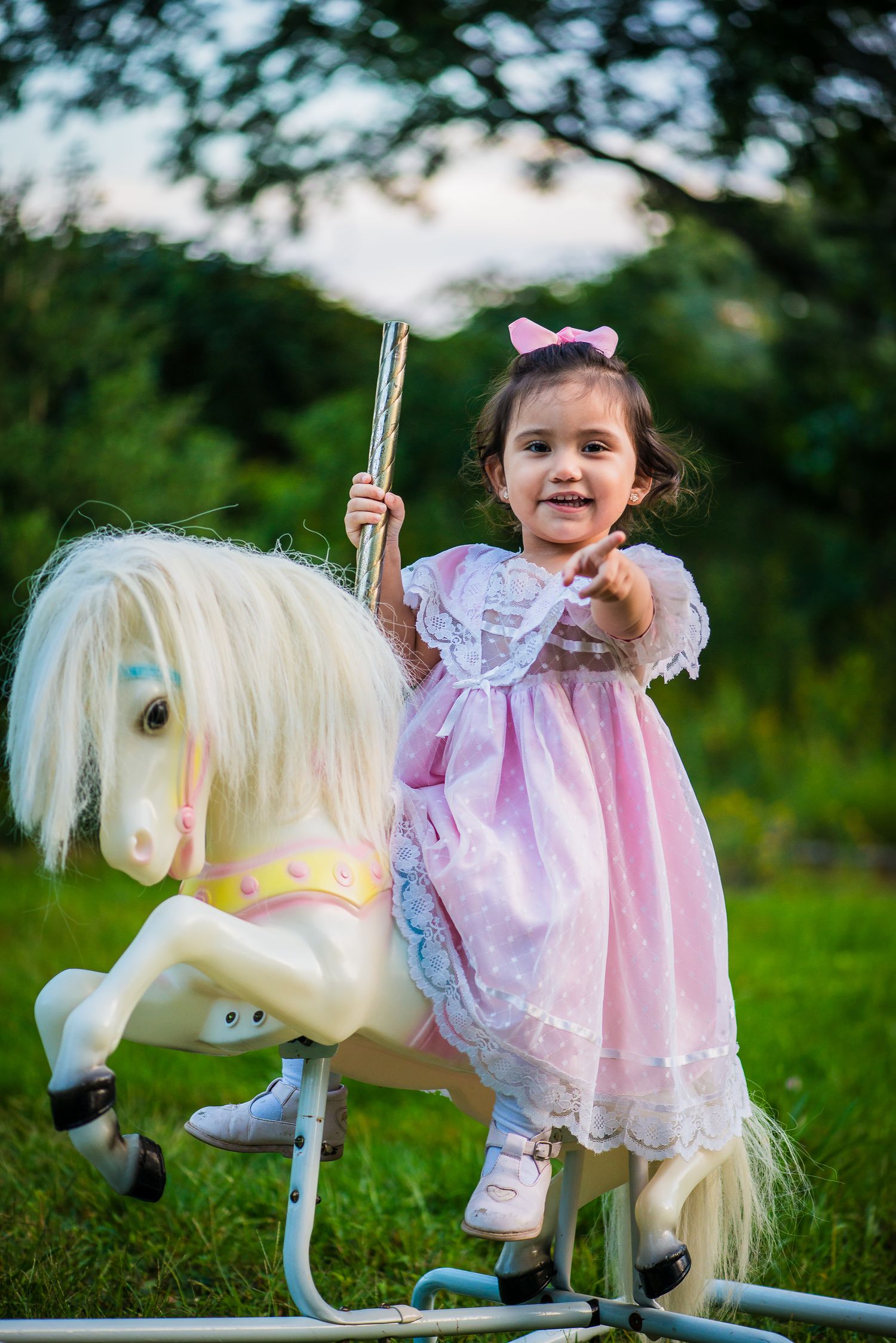 Happy toddler girl in pink dress riding white rocking horse toy in green outdoor setting.