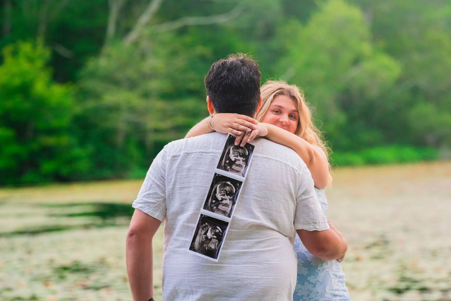 Happy couple embracing outdoors holding ultrasound photos, announcing pregnancy near tranquil lake.