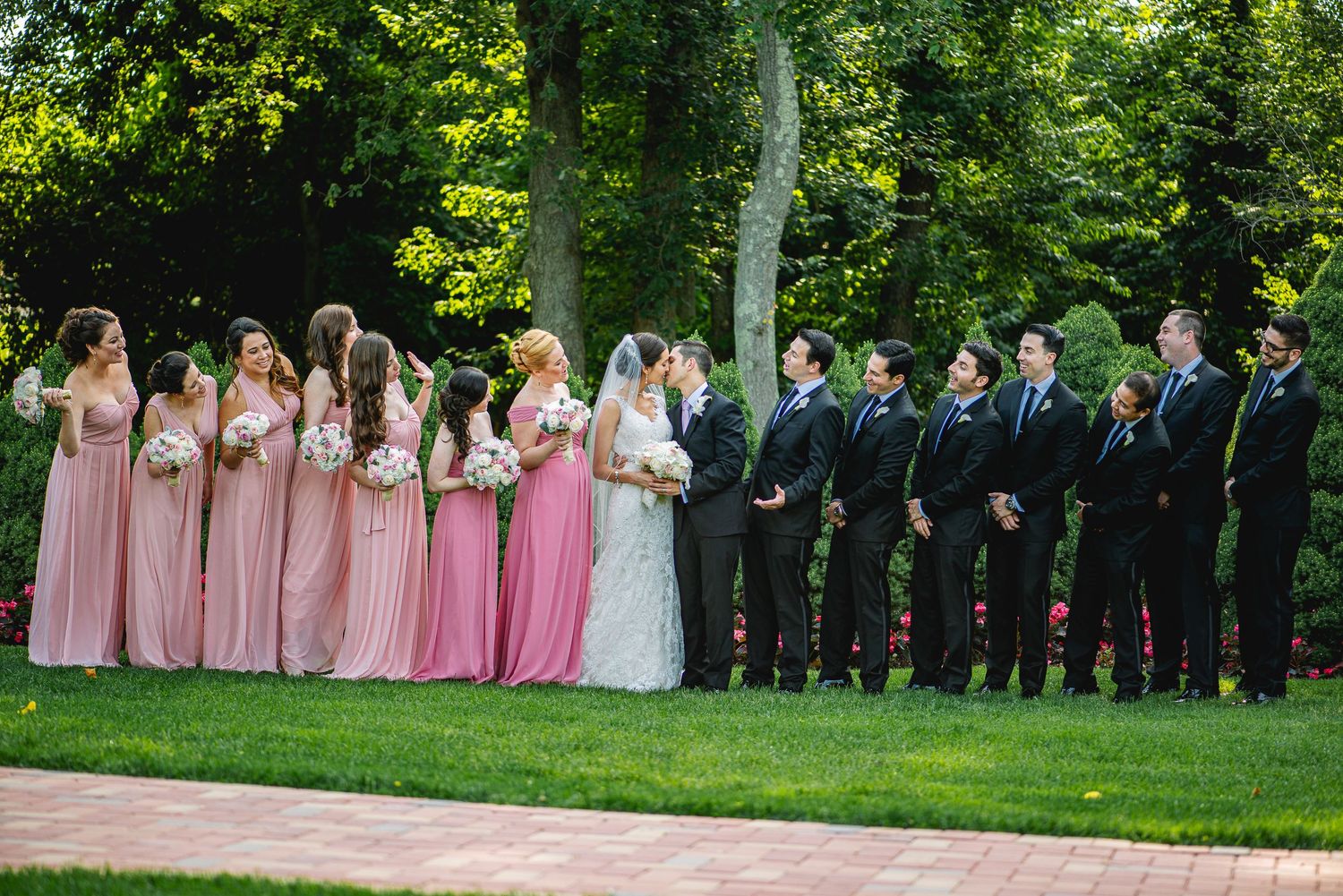 Wedding party poses outdoors with bridesmaids in pink gowns and groomsmen in black suits on a lush green lawn.