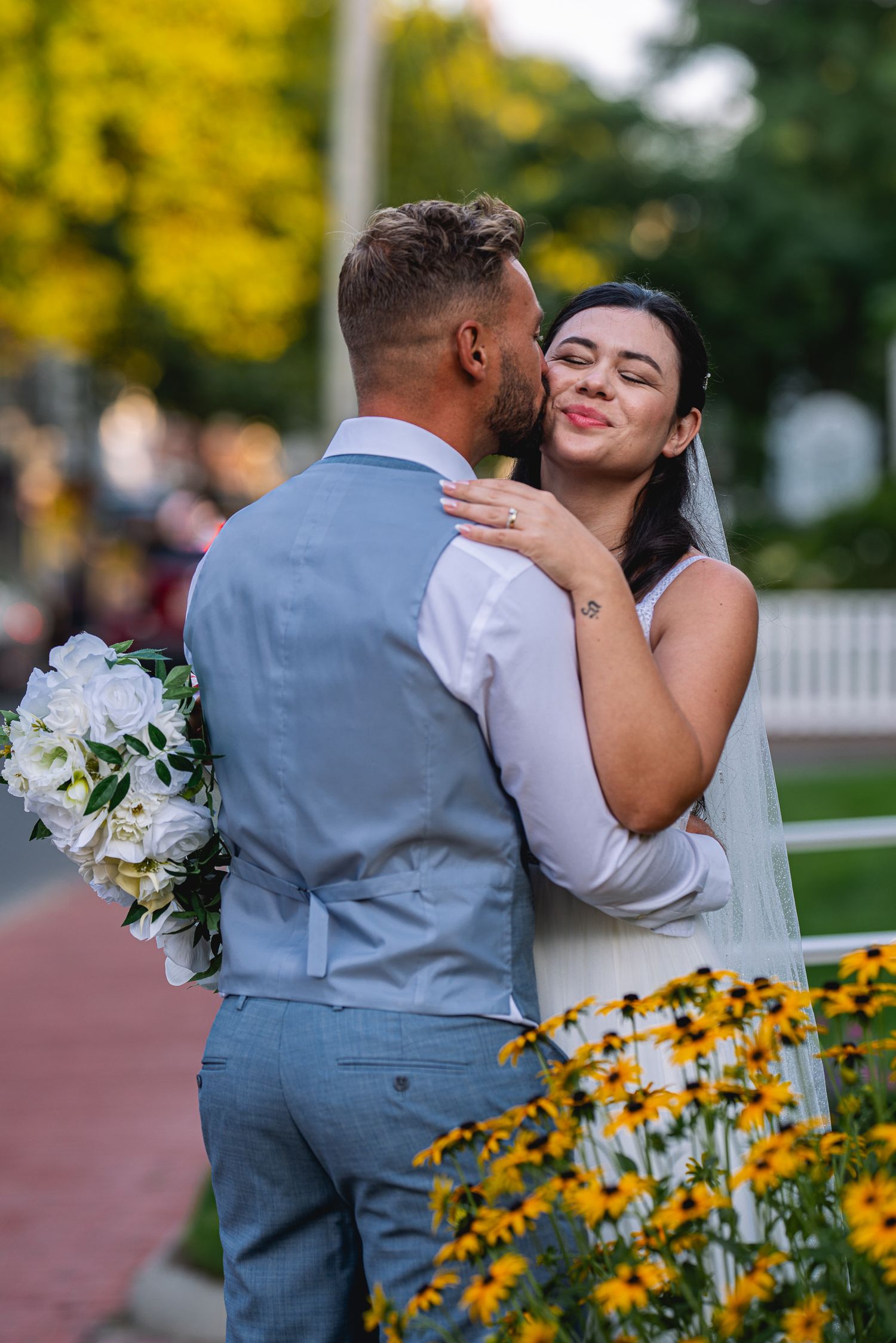 A groom in a gray suit kisses his smiling bride on the cheek while holding white roses outdoors.
