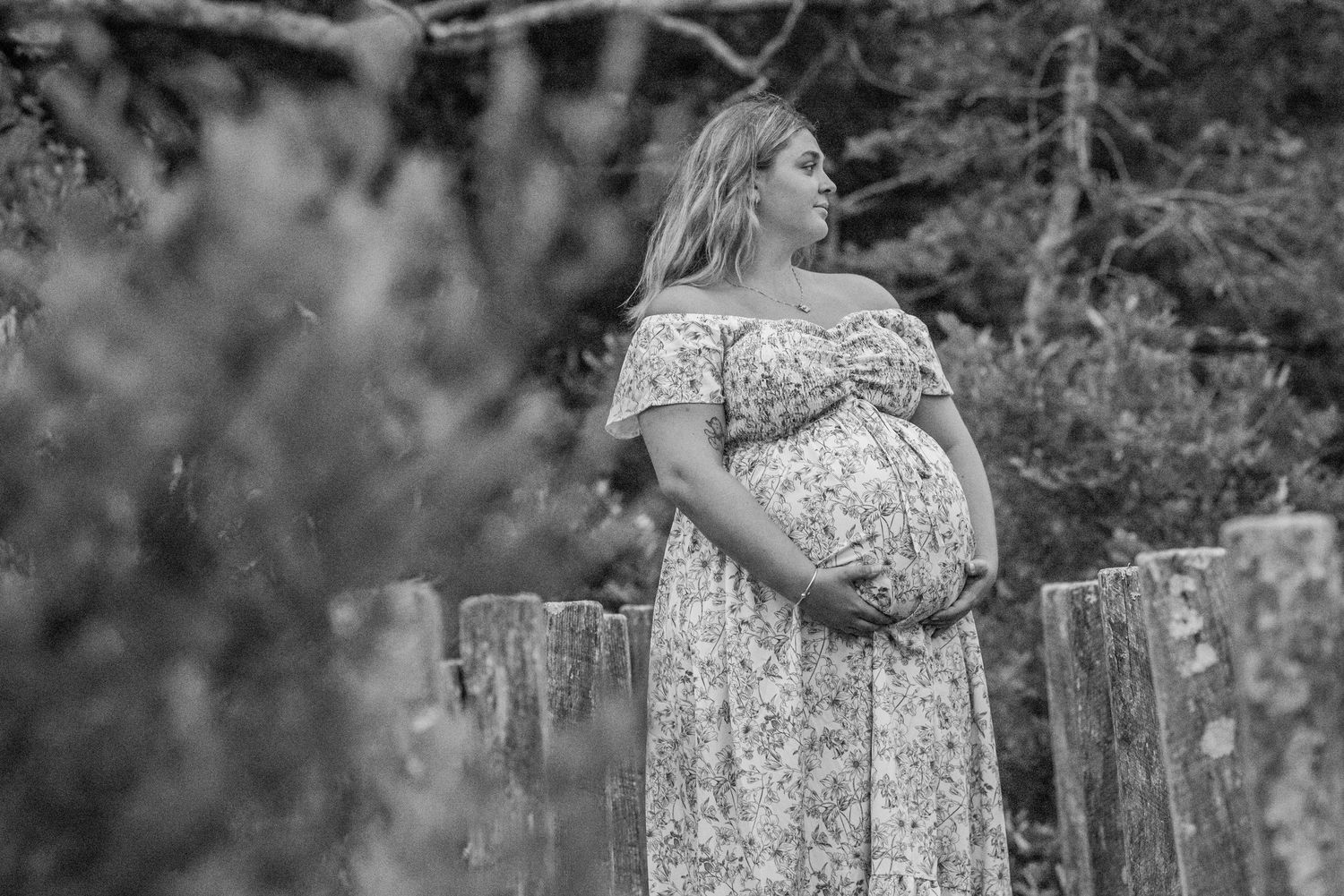 Black and white photo of a pregnant woman in a floral dress holding her belly outdoors near a wooden fence.