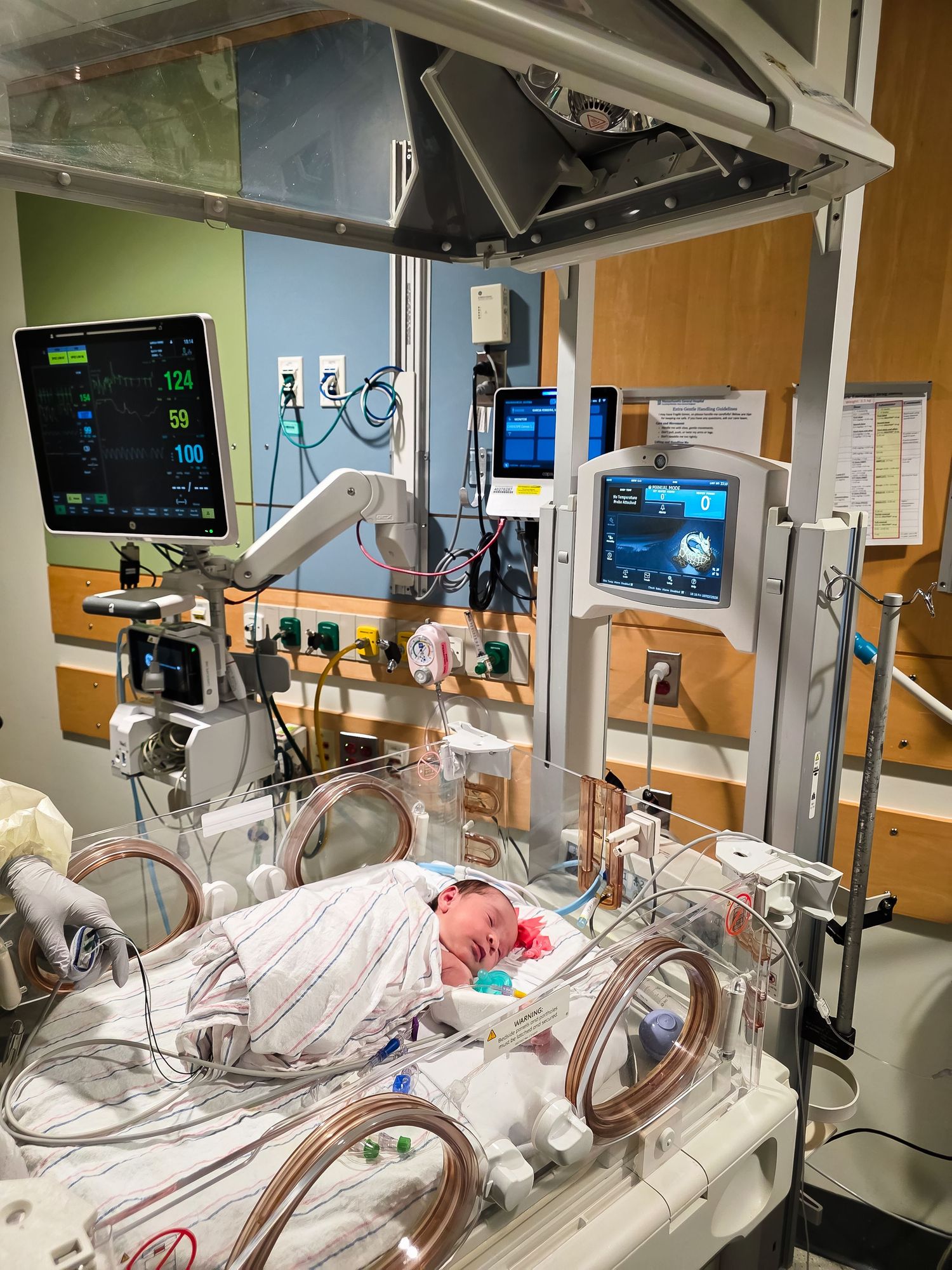 Newborn baby swaddled in a blanket sleeping inside a neonatal incubator in a NICU, surrounded by vital sign monitors and medical equipment.