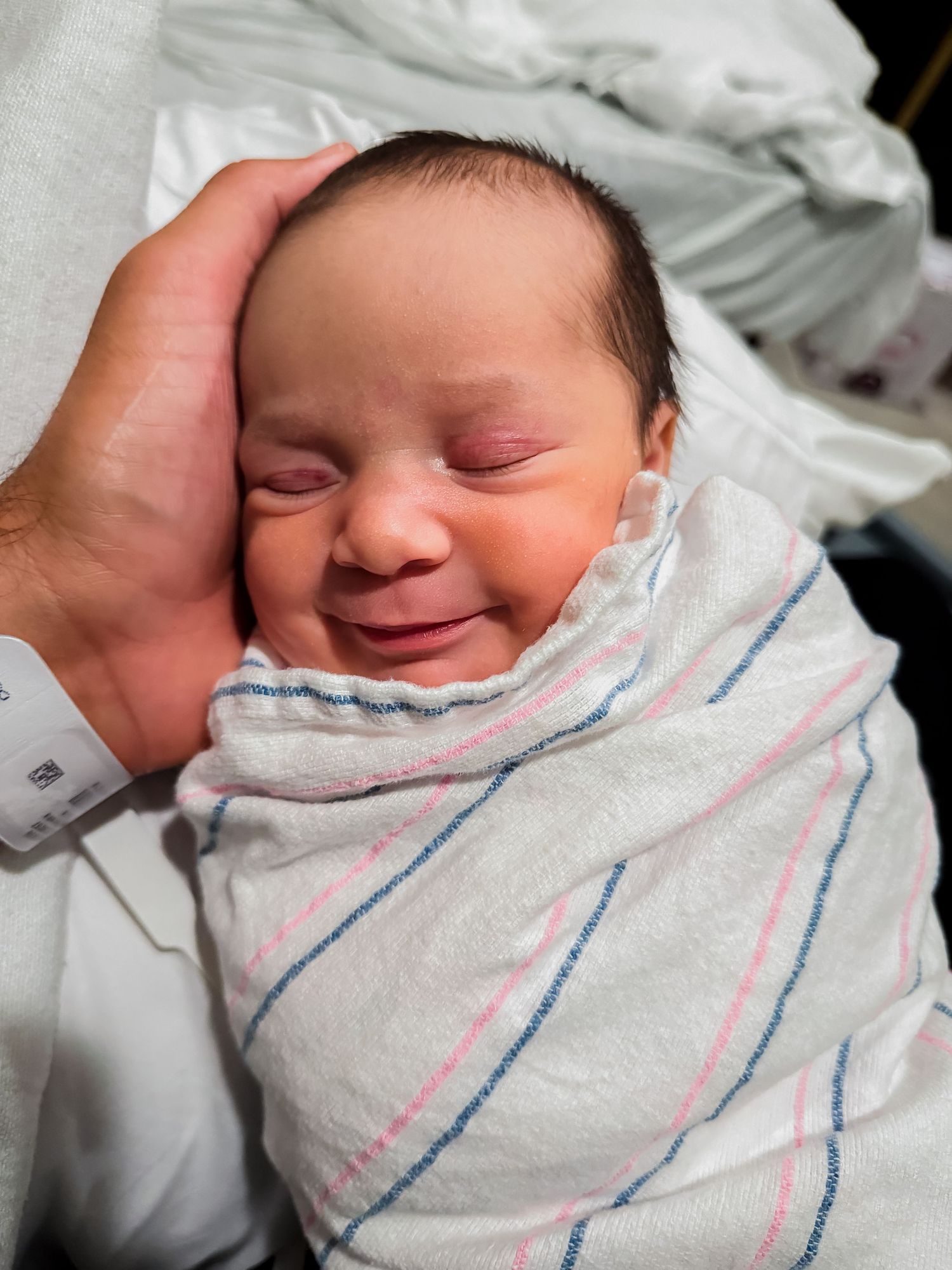 Smiling newborn baby with eyes closed, swaddled in a striped hospital blanket while a parent's hand gently cradles the head.