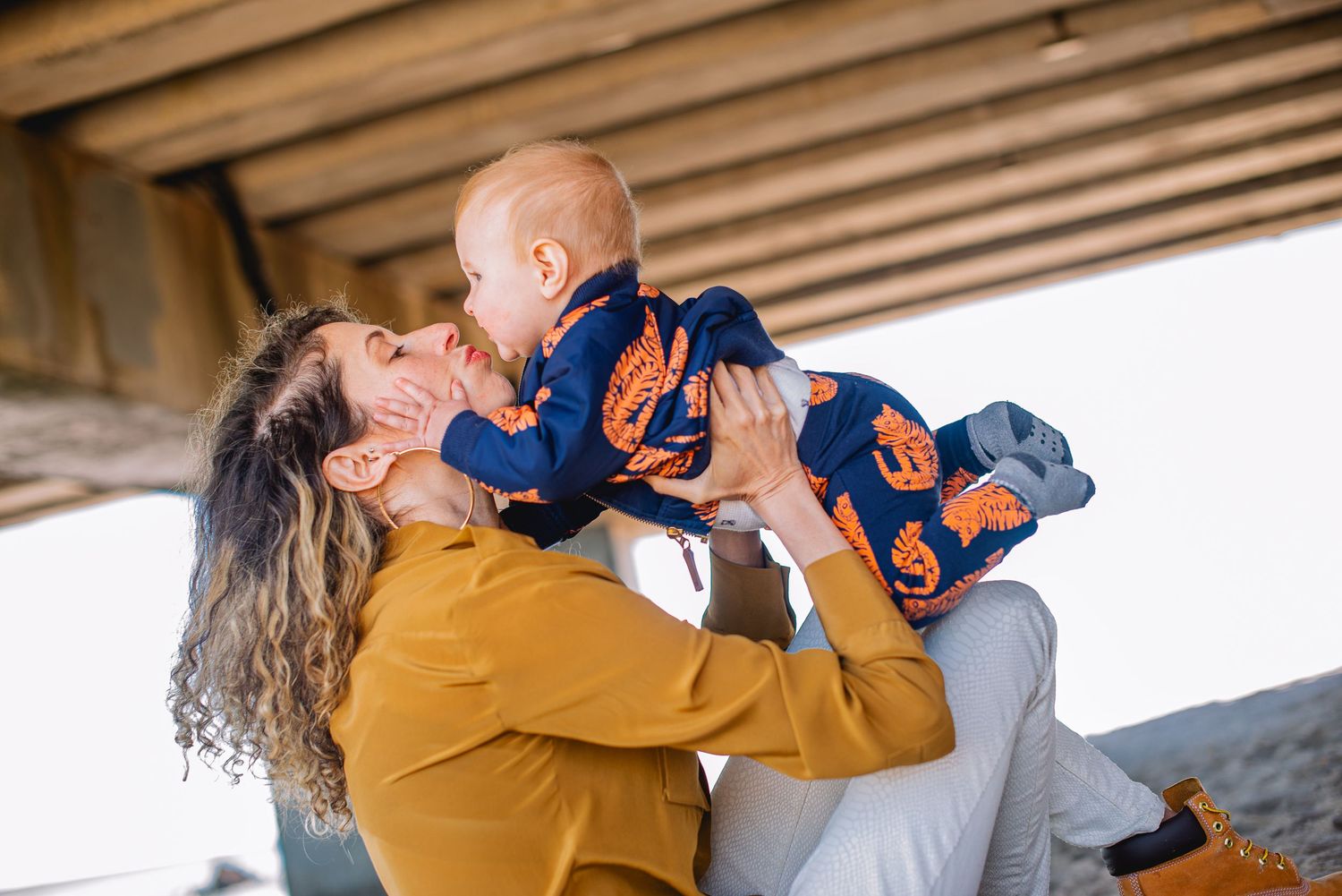 A joyful mother lifts her smiling baby under a wooden structure, both dressed in warm autumn colors.